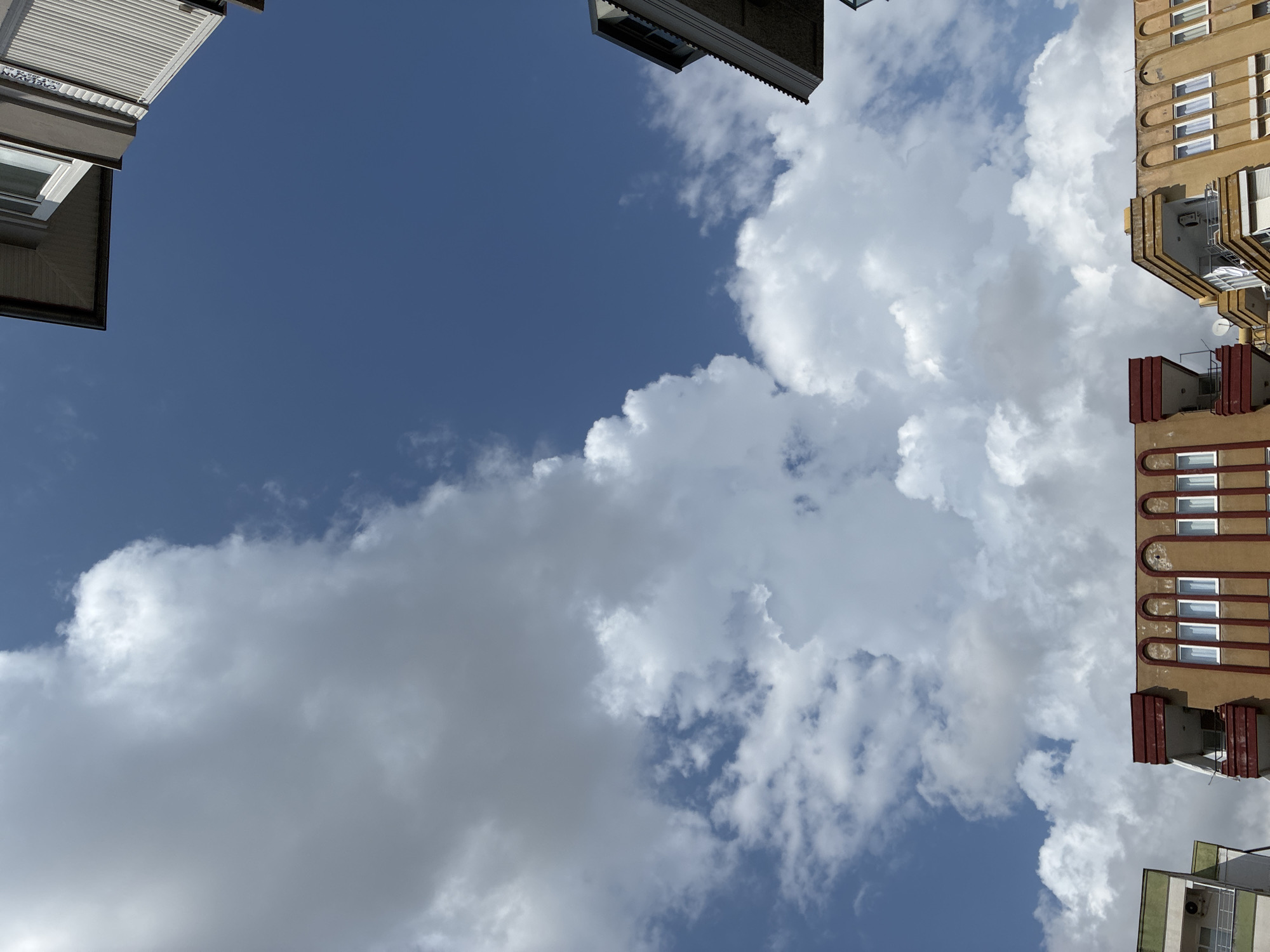 The image shows a view looking up at the sky from street level in Alanya, Turkey. The sky is a vibrant blue with scattered white cumulus clouds. The tops of several buildings are visible along the bottom and edges of the frame, suggesting an urban environment. These buildings appear to be residential, with balconies and windows. The architecture suggests a Mediterranean style. The image is oriented vertically, emphasizing the expanse of the sky.