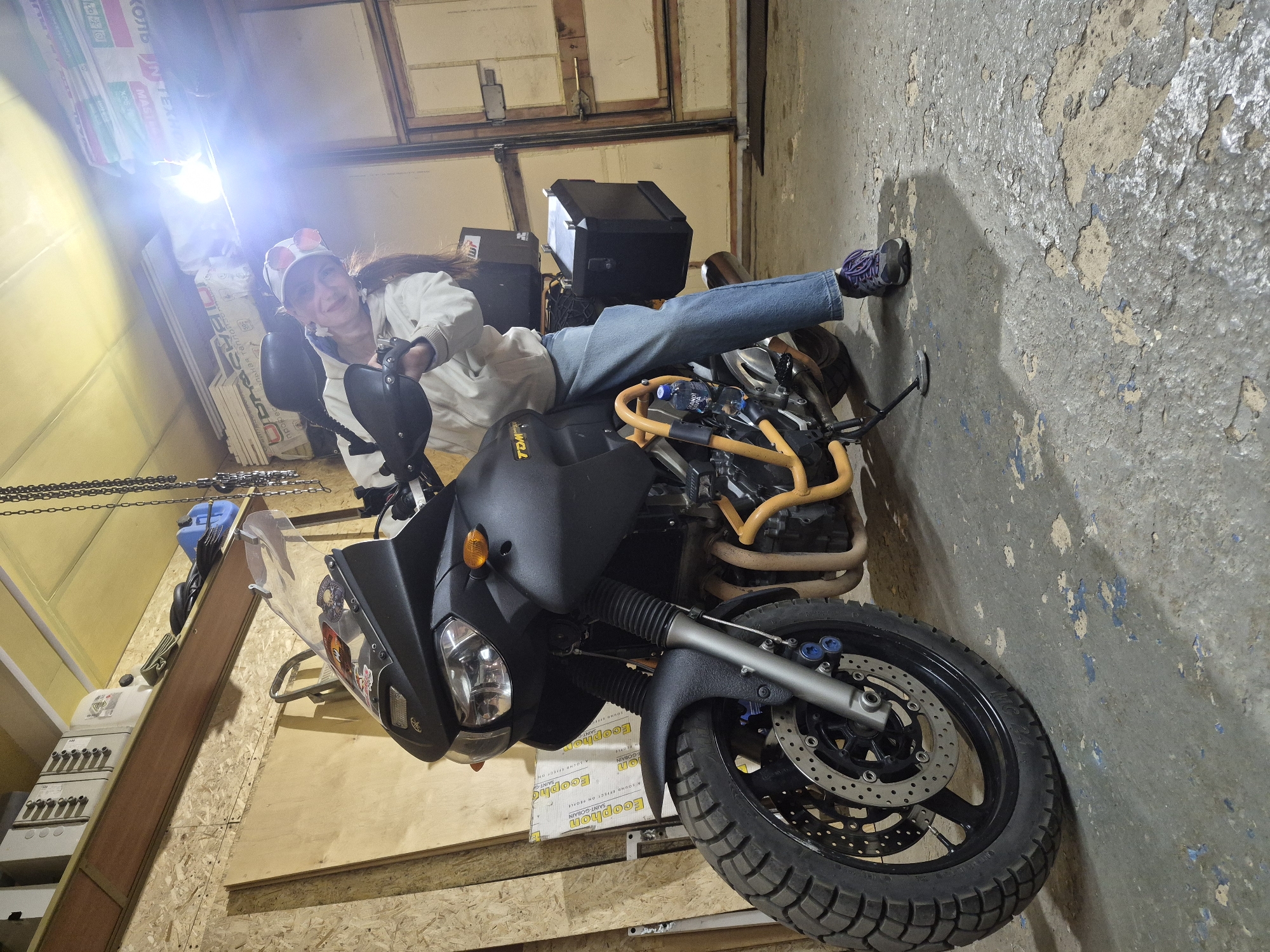 The photograph captures a woman reclining on a black motorcycle in what appears to be a garage setting. The woman is wearing a white jacket, jeans, and athletic shoes, and she has on a white cap. She is positioned on the motorcycle in a casual, relaxed manner.
The motorcycle is a prominent subject in the image, featuring a black matte finish, an orange crash bar, and saddlebags. The environment surrounding the motorcycle is a garage interior. The walls show signs of wear and tear, with peeling paint exposing the underlying material. The floor has a concrete-like texture. Stacks of packaging materials, labeled "Ecophon" and "Saint-Gobain" are visible.