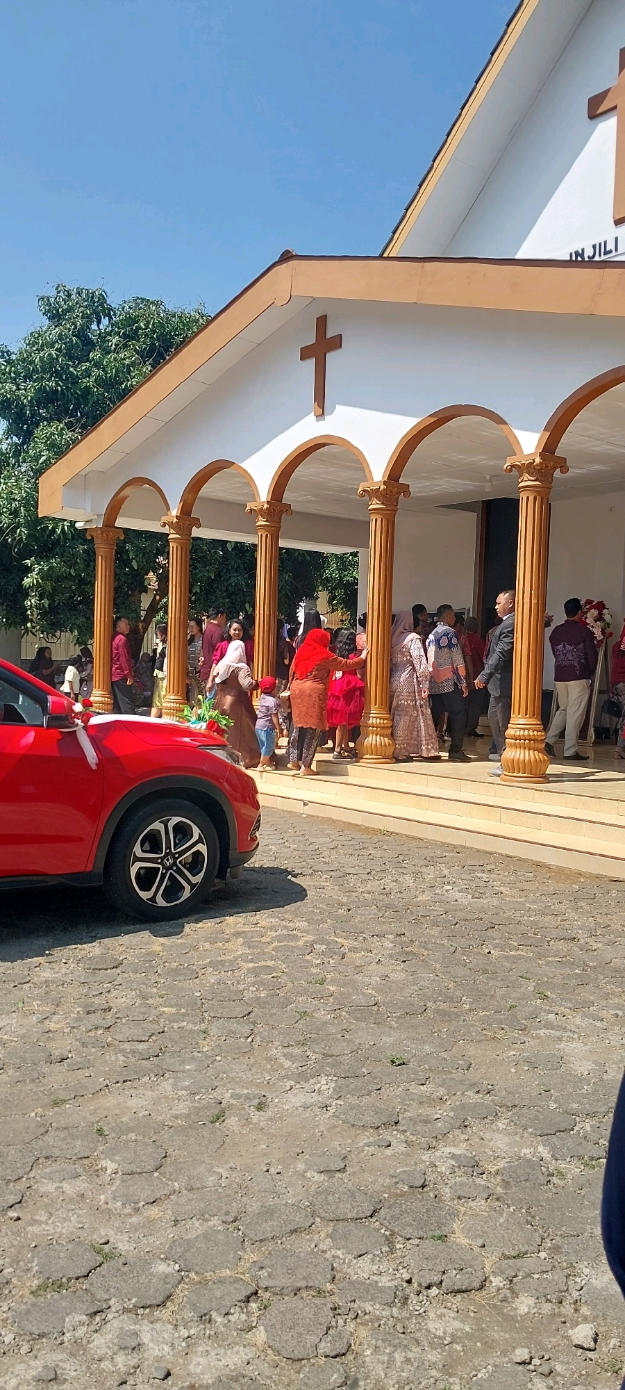 The image depicts the entrance of a church in Keling, Indonesia, on a sunny day. A red car decorated with a white ribbon and flowers is parked on a cobblestone surface to the left. A group of people, dressed in festive attire, are gathered on the steps and under the arched entryway of the church. Some are entering or exiting the building, suggesting a special occasion such as a wedding or religious ceremony. The church building has a white facade with brown trim and features prominent crosses. Trees are visible in the background.