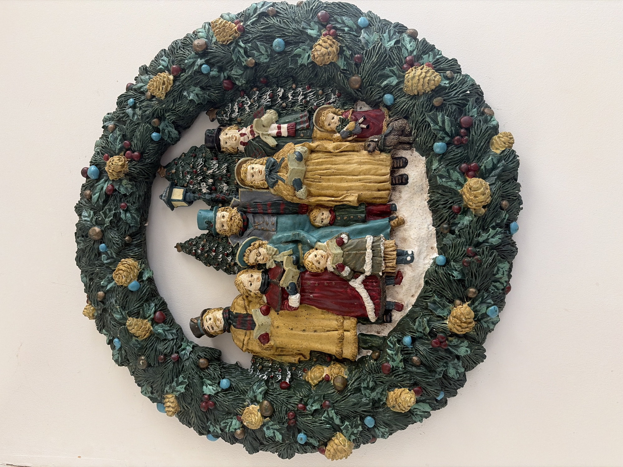 This media file features a highly detailed, three-dimensional Christmas wreath displayed against a plain white background, likely a wall. The circular wreath is crafted from sculpted material, possibly resin, and painted in rich, festive colors. The main body of the wreath is dark green, mimicking evergreen foliage, and is abundantly decorated with golden-brown pinecones, clusters of small red berries, and interspersed light blue/teal spherical ornaments.
Within the wreath's open center, a charming winter scene unfolds. A group of stylized figures, appearing to be people from a bygone era (possibly Victorian carolers), are arranged vertically. They wear warm, layered winter clothing, including long coats in shades of gold, red, blue, and olive green, complemented by hats, bonnets, and scarves. Several figures are depicted holding small, open books, suggesting they are singing carols. One smaller figure holds what appears to be a doll. The background of this inner scene includes miniature evergreen trees lightly dusted with white, sculpted snow, which also covers the ground around the figures.
The figures have simple, serene expressions, contributing to a nostalgic and festive atmosphere. The overall piece evokes a traditional Christmas or winter holiday gathering. There is no visible text in the image. The location context for this media file is Chatham, United Kingdom.