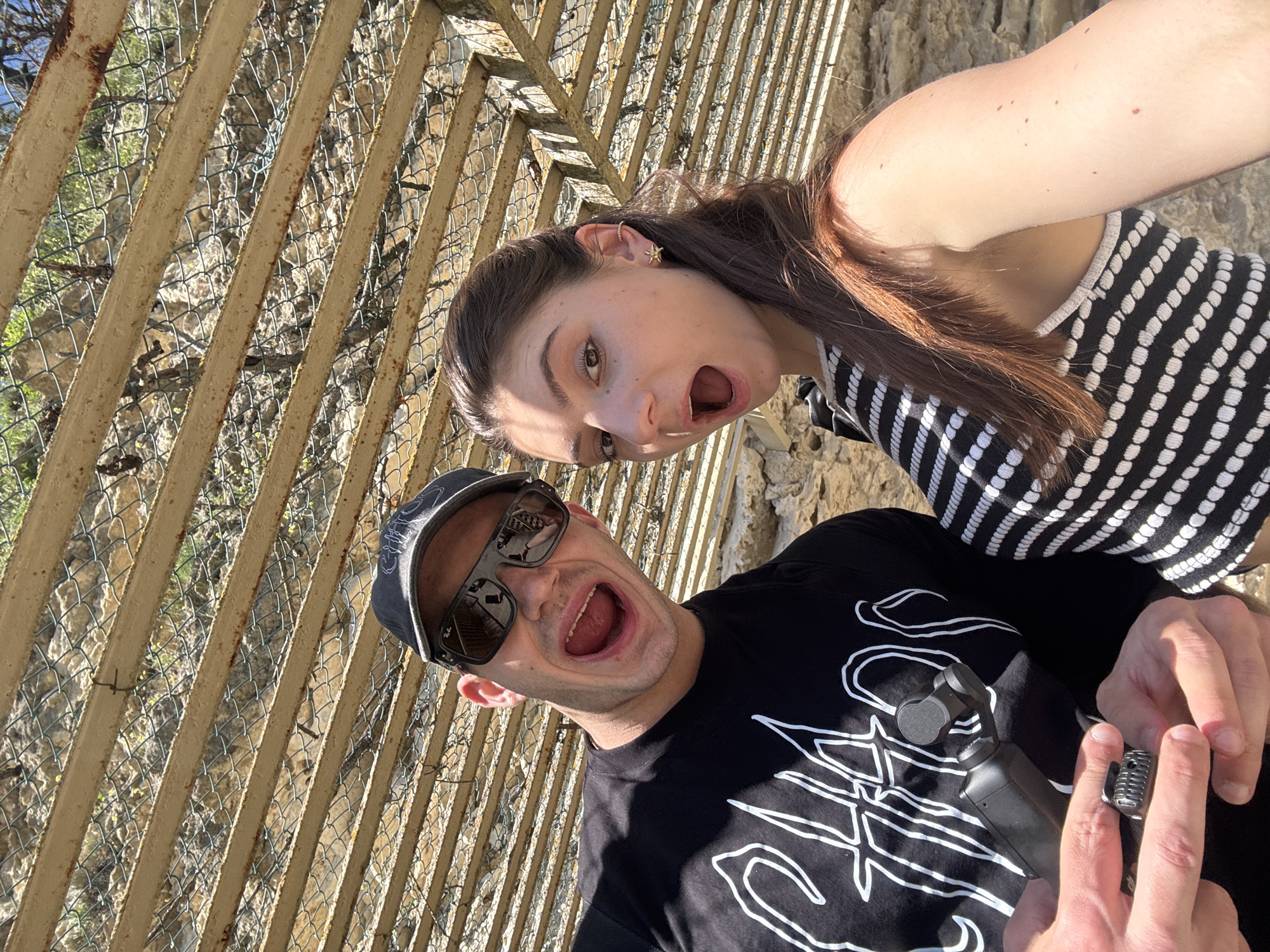A man and a woman are posing with their mouths open for a selfie in front of a metal fence and rock wall. The man is wearing a black t-shirt, a baseball cap, and sunglasses. The woman is wearing a black and white striped top. The man is holding a small black camera in his hands. The location is Unknown, Unknown.