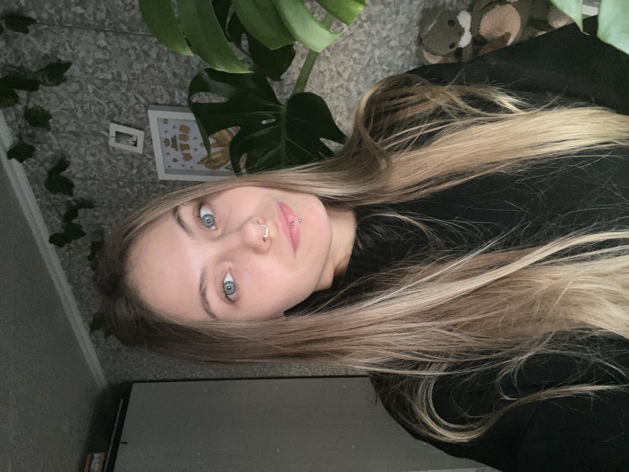 A woman with light brown and blonde hair, blue eyes, a nose ring, and lip piercings is facing the camera. She is wearing a black sweater. To her left are a green plant (possibly a Monstera) and a picture frame on a textured gray surface. To her right is a stuffed animal toy. The bottom of the frame shows a gray object. The location is likely indoors, but not specified as Фоки, Russia.