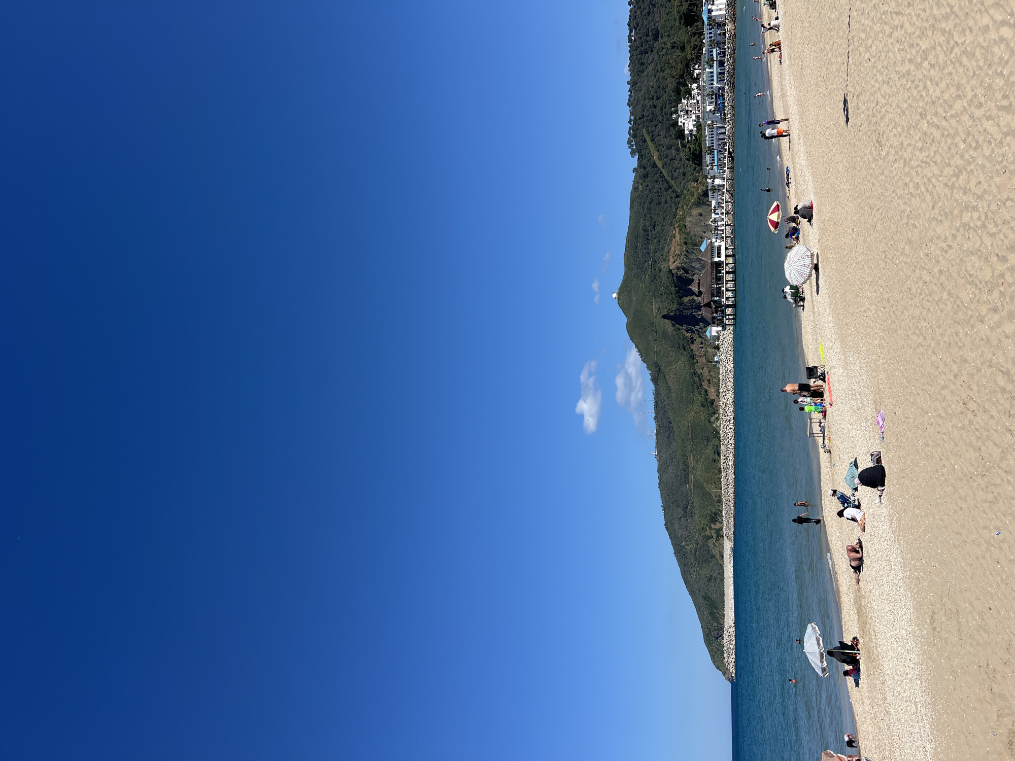 The image depicts a sunlit beach along the coastline of M'Diq, Morocco. The right side of the image features a beach populated with people engaging in activities like sunbathing, swimming, and relaxing under umbrellas. The beach is sandy, with a visible shoreline where the water meets the sand. The backdrop includes a line of buildings and residences adjacent to a steep, green hillside dotted with trees and vegetation. To the left of the image, the sky is a clear, deep blue, providing a strong contrast to the land and sea.