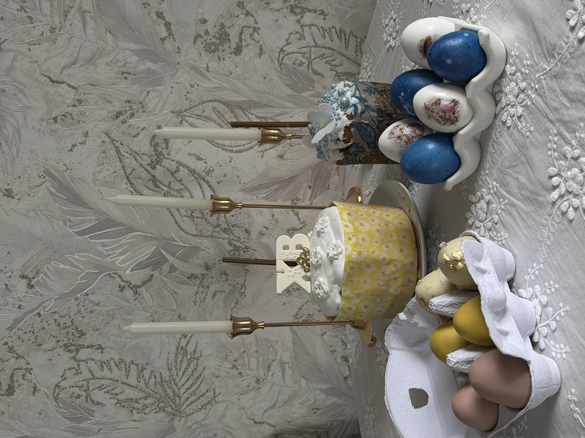 The image shows a still life arrangement. In the foreground, there's an open, white carton holding a mix of colored eggs: some brown, some yellow, and others with a cream color and gold leaf detail. Behind this, there's a cake decorated with a yellow and white daisy pattern, placed on a saucer. Behind the cake stands a white topper that reads "XB" in Cyrillic letters. To the left of the cake, there's a gold-colored candelabra holding three white candles. Behind the candelabra, there's another decorated cake, this one with blue floral patterns and sugar decorations. Behind that, there's a white egg holder containing several painted eggs, including blue and decorated ones. The background consists of a textured wall with a floral or feather-like pattern. The setting appears to be indoors, possibly in a home in Reshetnikovo, Russia, given the Cyrillic letters. The overall impression is of a celebratory or holiday table setting.