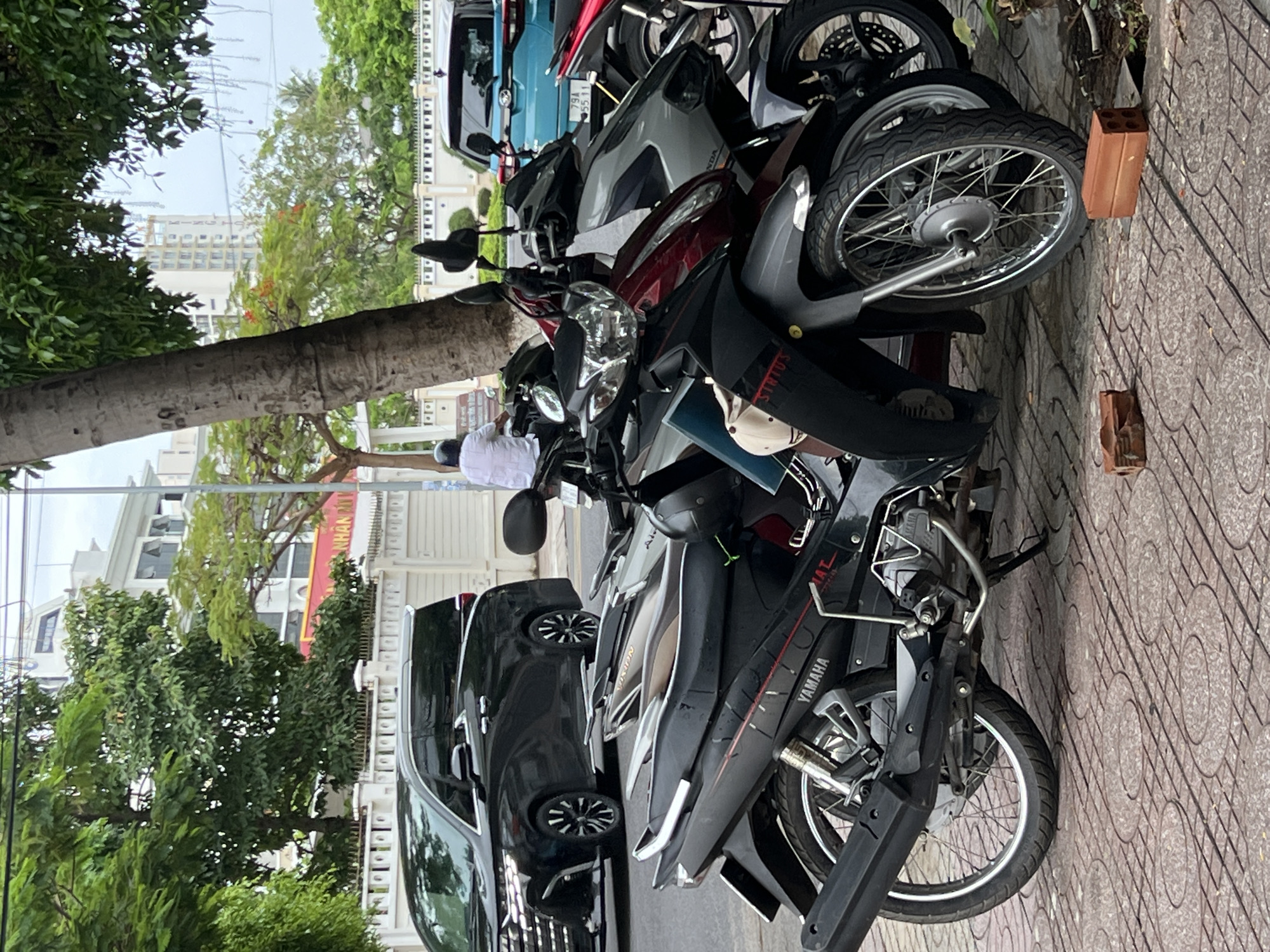 The image depicts a street scene in Bac Nha Trang Ward, Vietnam. In the foreground, several parked motorbikes dominate the view. A black car is parked alongside the motorbikes. In the background, a person in white clothing is walking on the sidewalk. Trees line the street and a building is visible. The pavement is patterned.