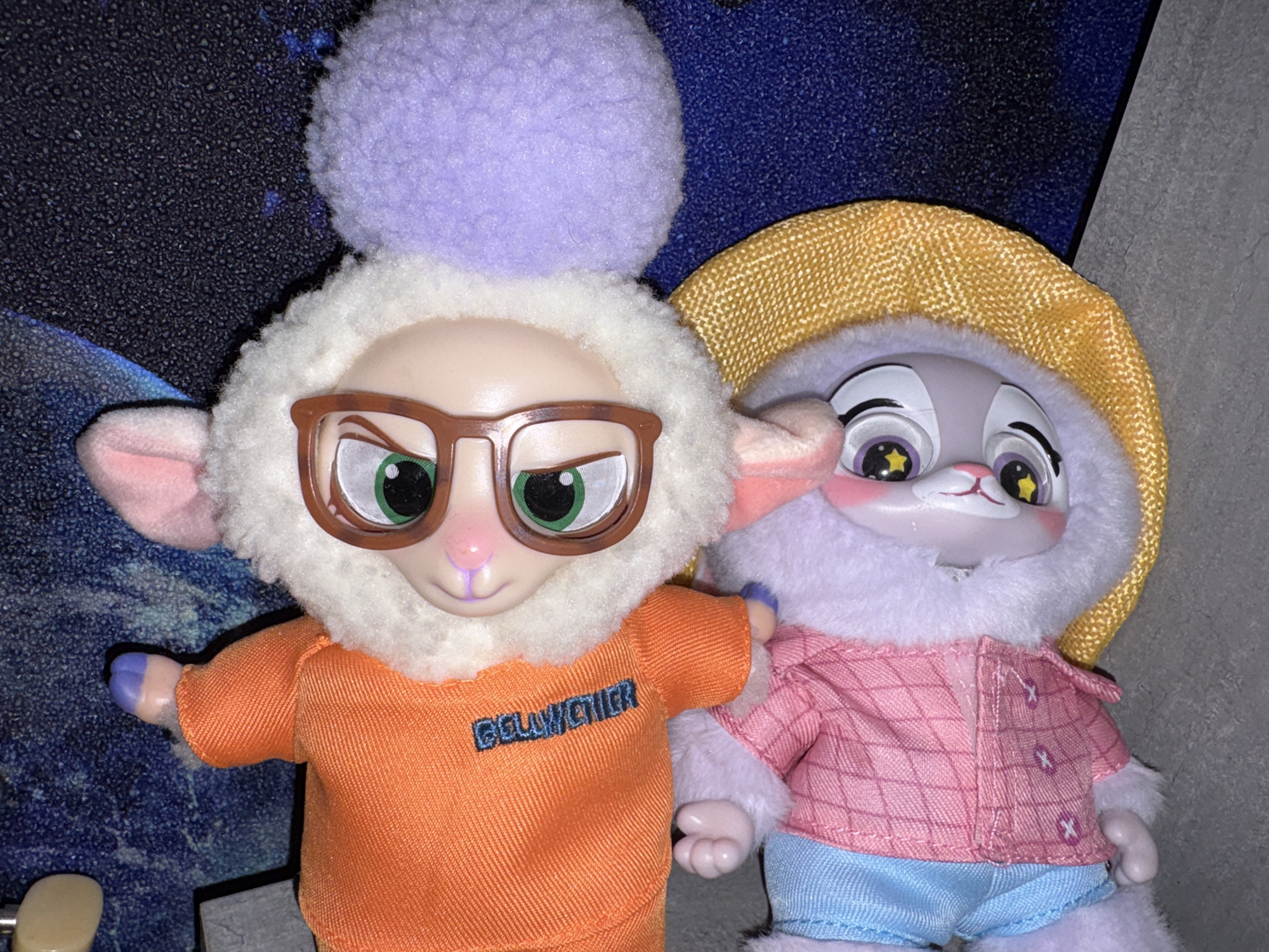 Two plush dolls are positioned side by side. On the left is a sheep doll wearing glasses, an orange shirt with "BELLWETHER" printed on it, and a purple pom-pom on top of its head. To the right is a rabbit doll wearing a yellow hood, a pink plaid shirt, and blue shorts. The background is dark and textured, possibly a wall or fabric. The location is specified as Прикамский, Russia, but there are no visual cues to confirm this location; it's just a context detail.