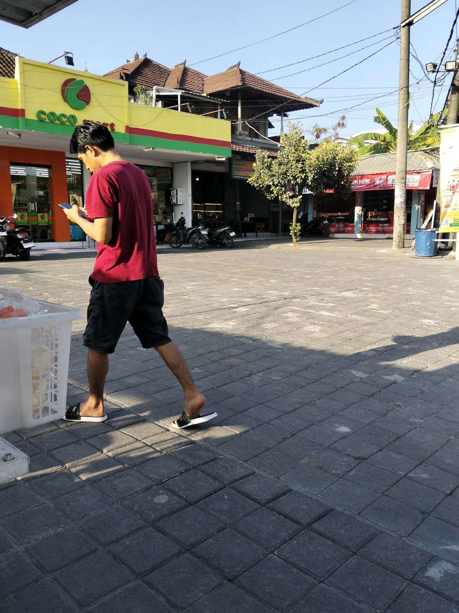 A young man wearing a red t-shirt and black shorts walks across a paved outdoor area, looking at his mobile phone. He is wearing black slides on his feet. To his left, a white plastic crate is partially visible. In the background, a building with a yellow facade and red and green trim is visible, featuring a "COCO MART" logo. The building appears to be a shop. Further in the background, there are more buildings, trees, and a blue trash bin. The scene is illuminated by sunlight, casting shadows on the pavement. The setting is consistent with a street scene in Kuta Selatan, Indonesia, likely near a commercial establishment.
