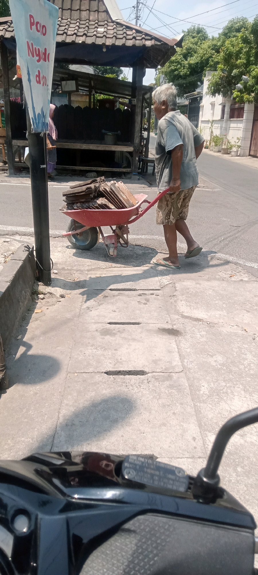 An elderly Indonesian man with gray hair is pushing a red wheelbarrow filled with roof tiles along a sidewalk in Surakarta, Indonesia. He is wearing a light gray t-shirt and patterned shorts, and flip-flops. The man is walking away from the camera, towards a small roadside stall with a tiled roof. A woman in a pink headscarf is visible inside the stall. In the foreground, a portion of a black motorcycle is visible, indicating the photo was taken from its perspective. The scene depicts a typical street in Surakarta, with concrete sidewalks, asphalt roads, and buildings in the background. The weather appears to be sunny with shadows cast on the ground.