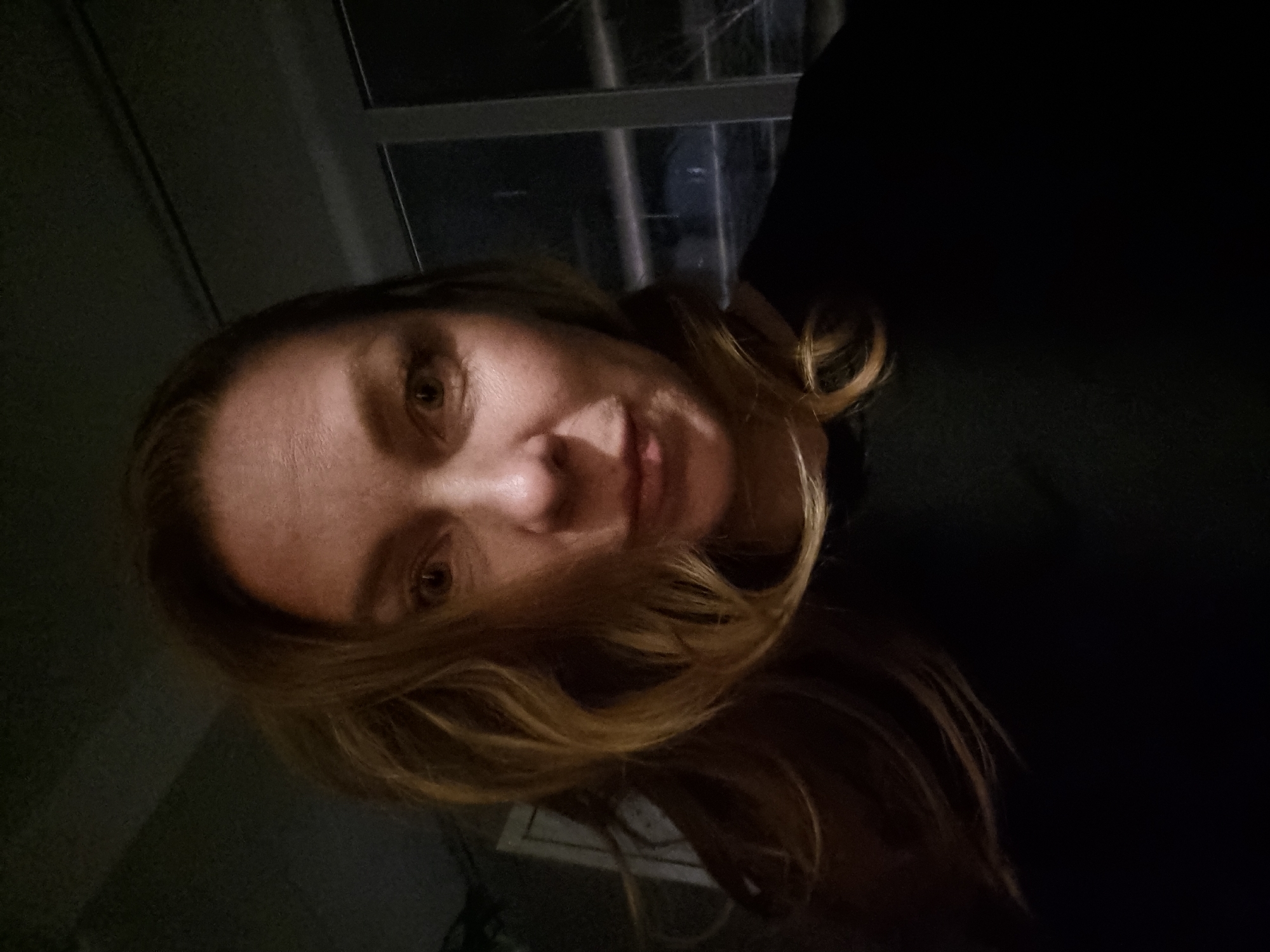 A woman with blonde hair is in an indoor environment. She is wearing a dark-colored garment. A window is visible in the background. The image is taken at an oblique angle. The location is not determinable.