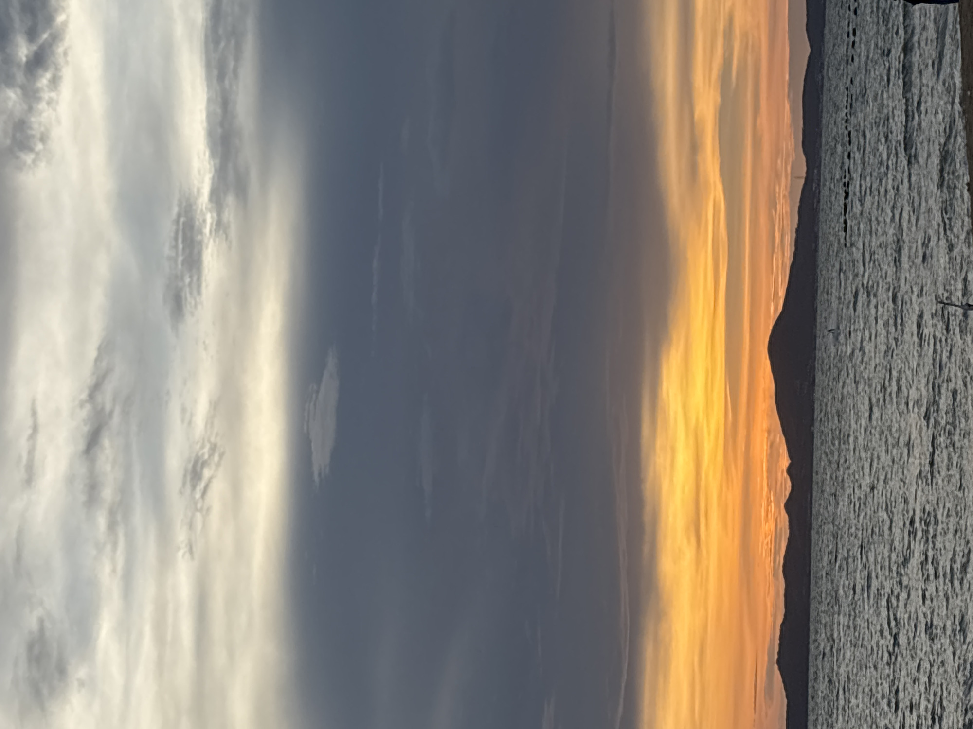An evening ocean view in Pattaya City, Thailand, features a dramatic sky with layers of clouds. The upper sky is a pale gray with streaks of white, transitioning to a darker gray layer. Below this, the sky ignites with a vibrant gradient of orange and yellow, characteristic of a sunset. In the foreground, the choppy surface of the ocean reflects the sky's colors. A silhouetted mountain range forms a dark line across the horizon, separating the sea from the sky. The scene is captured at eye-level, looking out over the water towards the distant landmass.