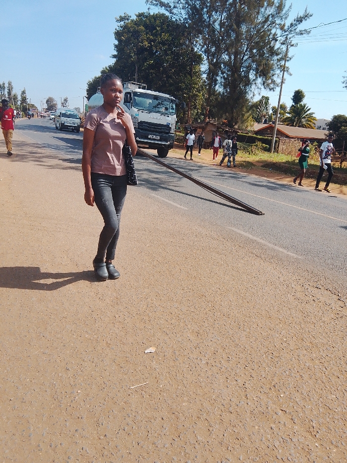 A young woman in a short-sleeved mauve shirt and dark jeans walks along a paved road, carrying a black purse. Behind her, a white tanker truck is parked near the side of the road, and several people are walking in the distance on the other side of the road. There is a long, dark pole that stretches across the width of the road. Trees and buildings are visible in the background. The sky is clear and blue.