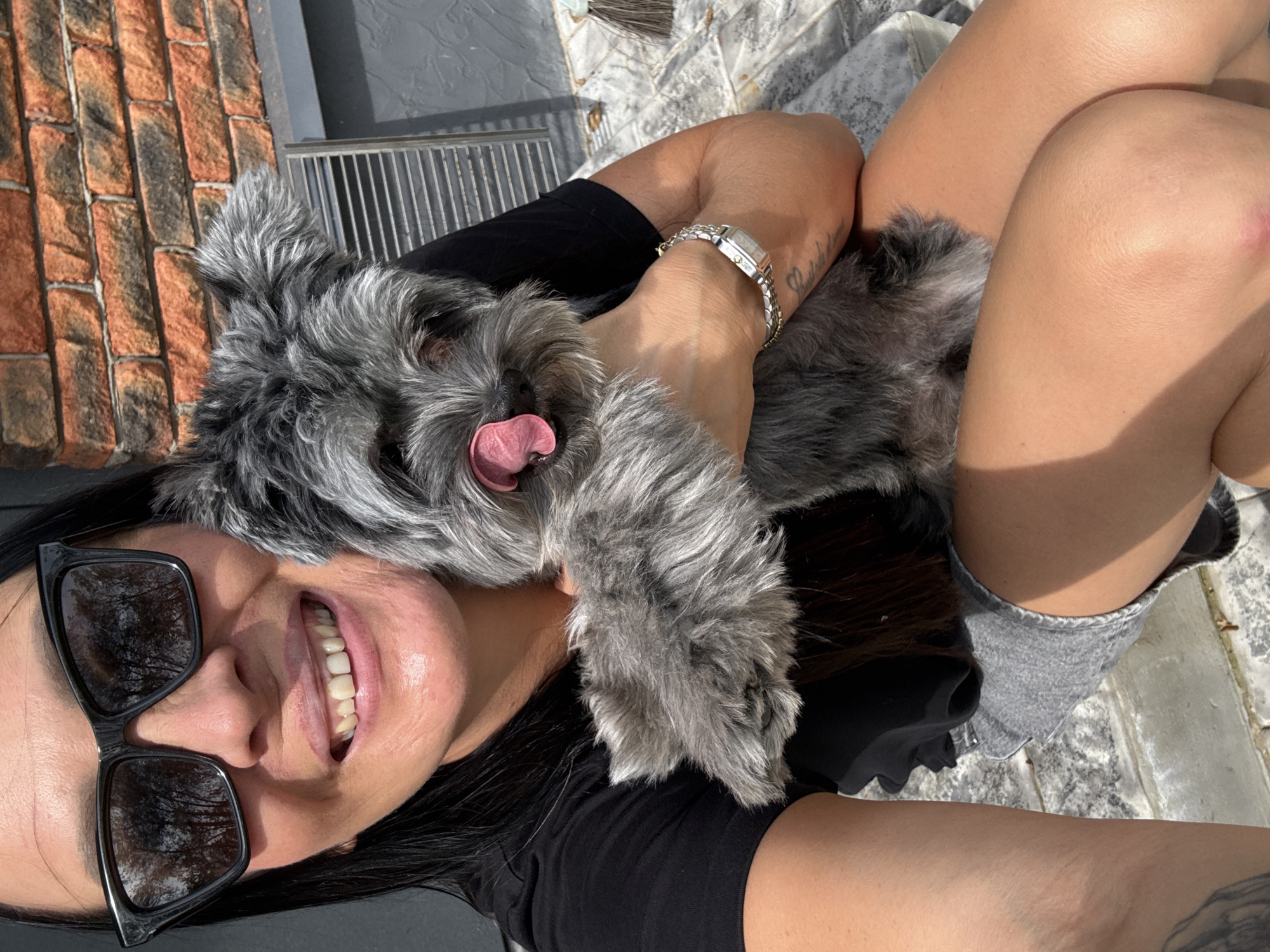 In a selfie-style shot, a woman with dark hair and sunglasses smiles broadly, holding a small, grey, fluffy dog. The dog has its tongue out and is looking towards the camera. The woman is wearing a black t-shirt and grey shorts. She has a watch on her left wrist. The background consists of a brick wall and a metal grate. The lighting suggests an outdoor setting, possibly a sunny day. The location is identified as Chaikovsky, Russia.