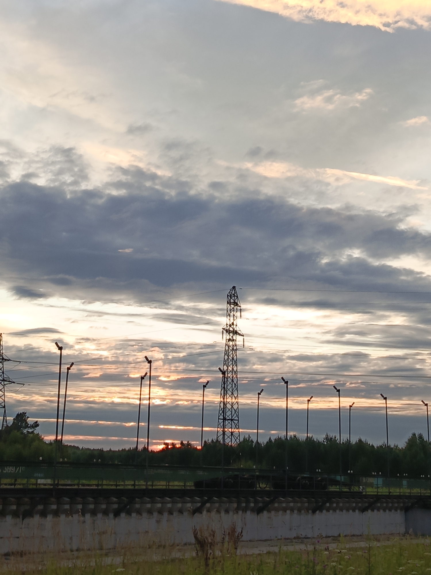 The image depicts a scene in Kauchuk Stantsiya, Russia, featuring a power pylon and a row of streetlights against a cloudy sky at sunset. The sky is filled with horizontally layered clouds, some illuminated with soft pink and orange hues from the setting sun, while others are dark gray. A tall lattice power pylon stands prominently on the left side of the frame, with power lines stretching across the sky. To the right of the pylon, a series of dark, slender streetlights with illuminated tops are evenly spaced. Below the sky, a dense line of green trees forms the horizon. In the foreground, a concrete structure runs horizontally across the bottom of the image, with a textured surface. Grass and some wildflowers are visible in the very bottom edge of the frame.