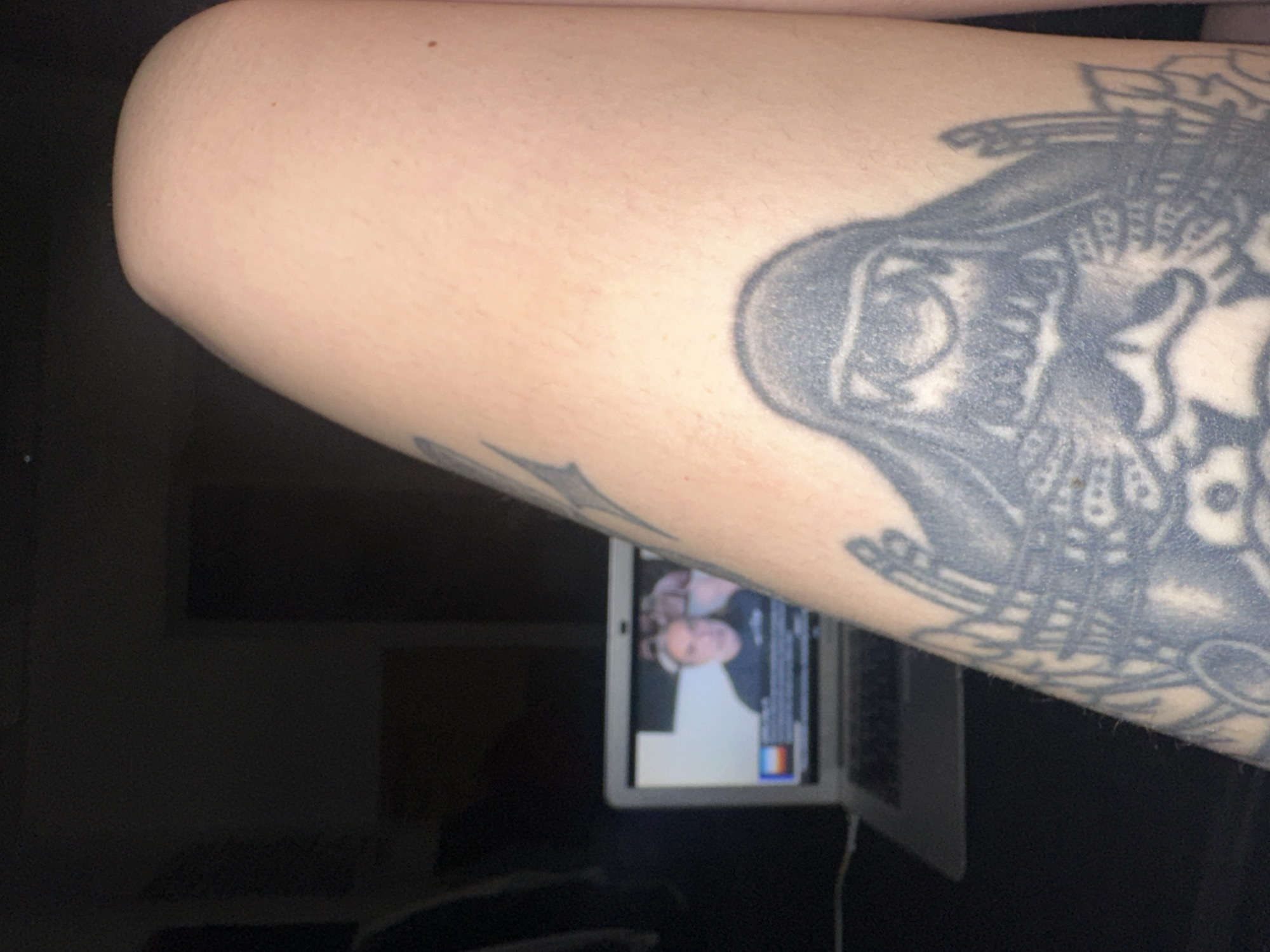 An arm with a tattoo is positioned in the upper portion of the image, to the right. The tattoo is black and gray and shows an intricate pattern of shapes and lines. Below the arm is a partially visible laptop. The screen of the laptop displays a person's face. The background is dark. The image's location is unknown, but it is unlikely to be in Buenos Aires, Argentina based on the objects in the photo.