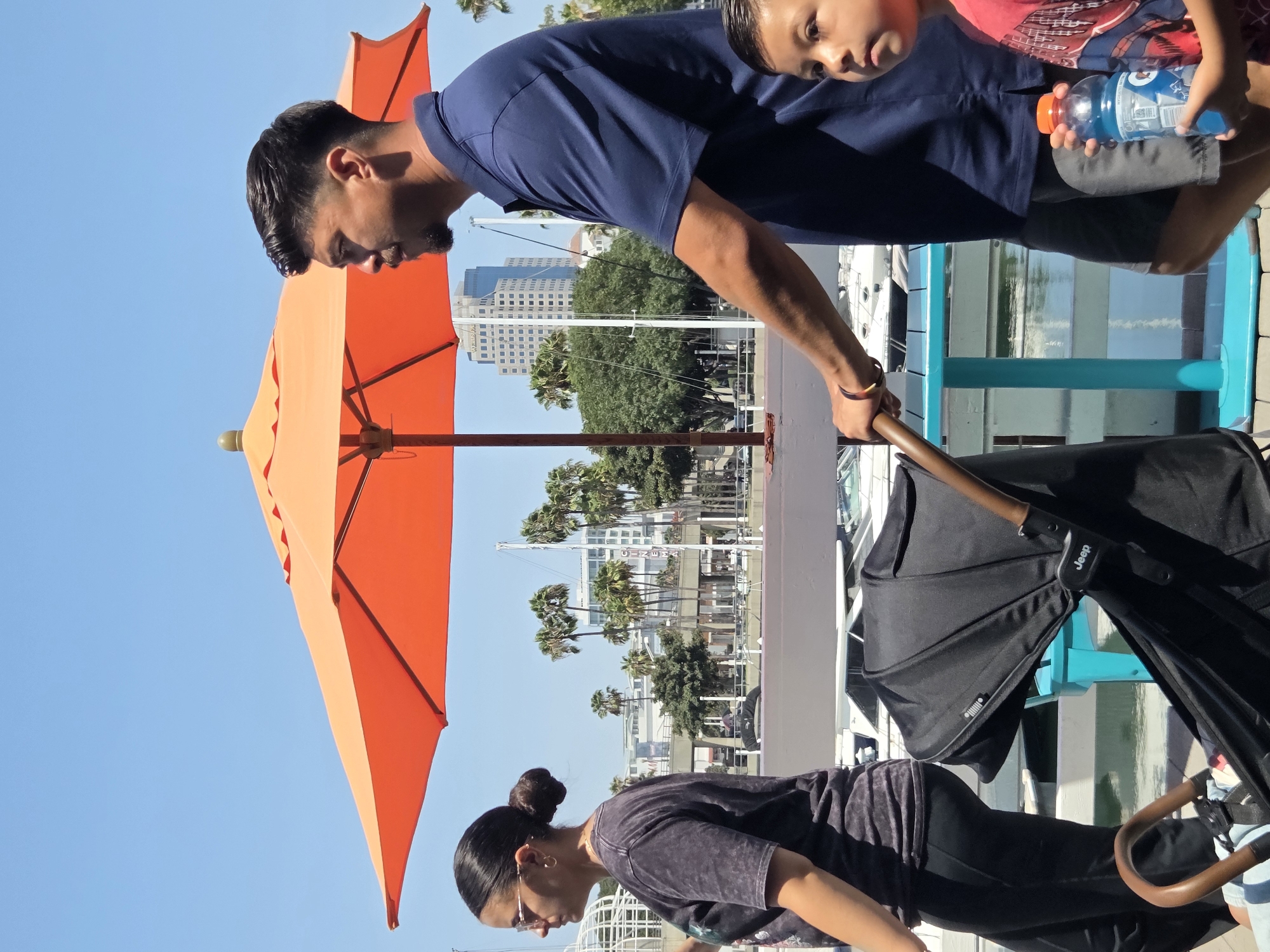 In Long Beach, California, under a clear blue sky, an orange umbrella casts a shadow over a waterfront scene. A man in a navy blue polo shirt and grey shorts walks with a stroller, his right hand gripping the handle. Beside him, a woman in a dark t-shirt and black pants walks, her hair pulled back in a bun, wearing glasses and hoop earrings. A young boy in a red and blue shirt stands to the man's right, holding a blue sports drink bottle. The stroller, a black Jeep model, has a wooden handle. In the background, palm trees and modern buildings dot the cityscape, with boats docked along the waterfront. The foreground is occupied by the stroller, a bright blue table, and the surrounding paved walkway.