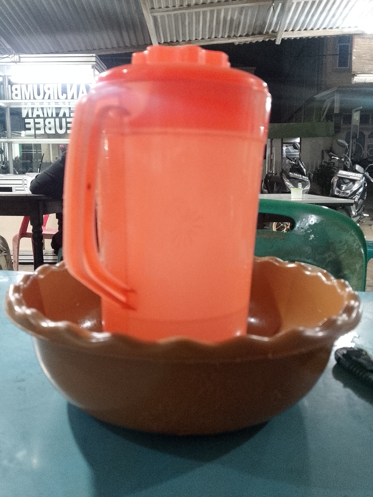 An orange plastic pitcher sits in a brown plastic bowl. The bowl rests on a blue table. In the background, there are motorcycles, chairs, and a sign with reversed lettering. The location is Lhokseumawe, Indonesia.