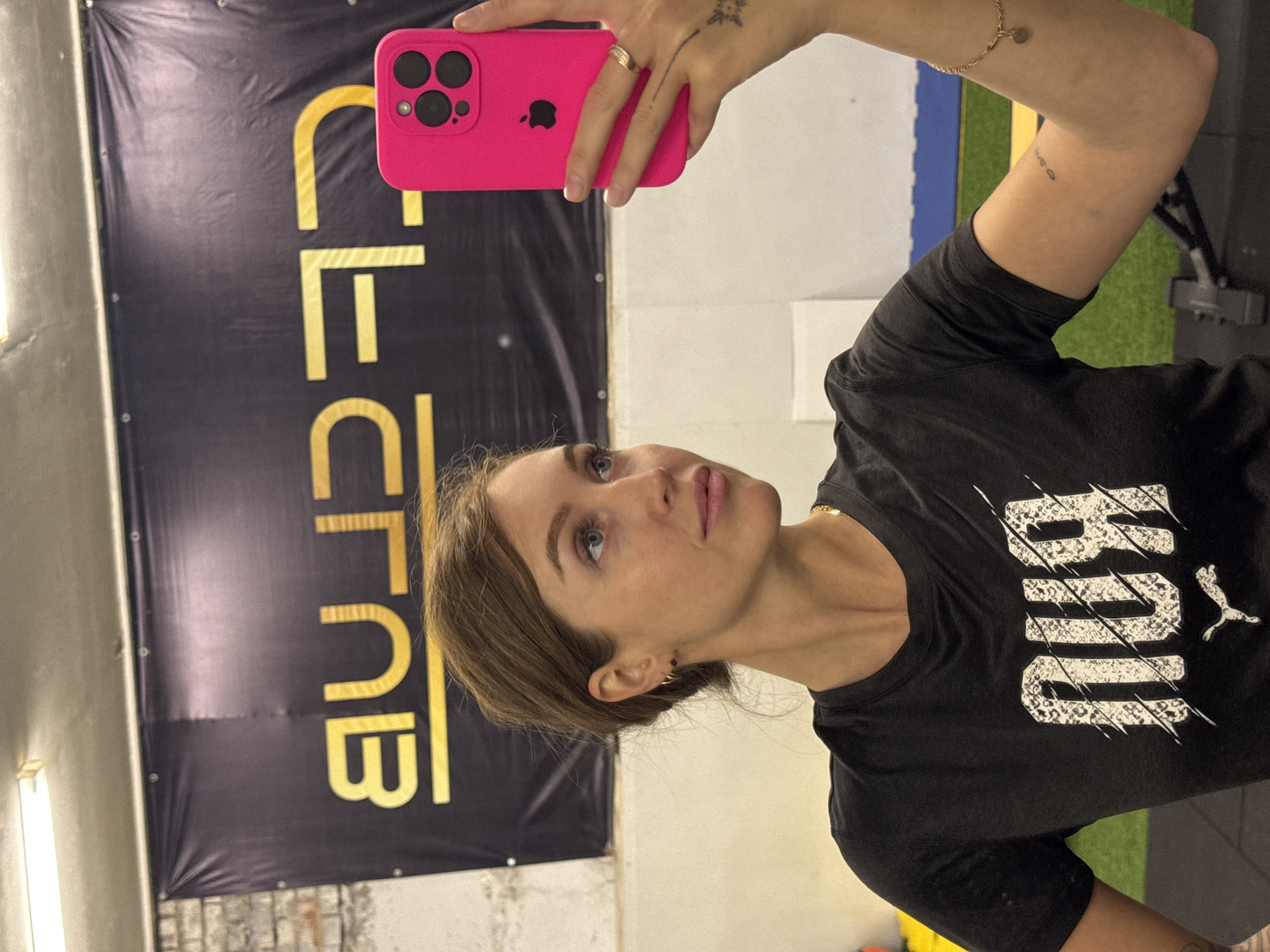 A woman with fair skin and shoulder-length brown hair is holding a pink smartphone in her right hand, taking a selfie. She is wearing a black Puma t-shirt and is positioned in what appears to be a gym or fitness studio. Behind her, there's a black banner with yellow lettering; the word "CLUB" is visible. The walls are partially visible, with some brickwork near the bottom of the banner. There's also gym equipment and flooring visible. The location of this image is not clear.