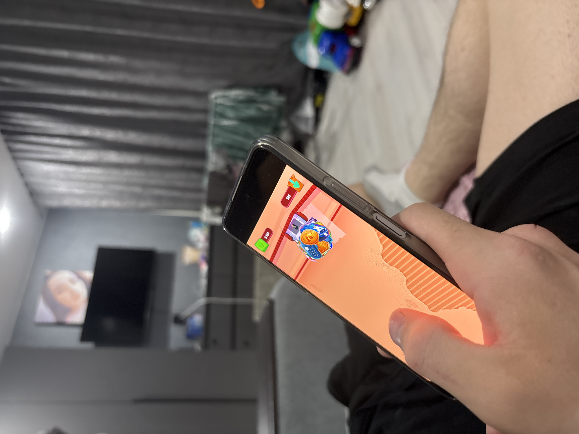 The image shows a person holding a smartphone, playing a mobile game. The game appears to be a casual puzzle or simulation type, with cartoonish graphics and a UI indicating progression or resources. The person's thumb is visible on the screen, likely interacting with the game. In the background is a bedroom interior, including a bed, curtains, and a wall-mounted television. There is no evidence that the image was taken in Chaikovsky, Russia.