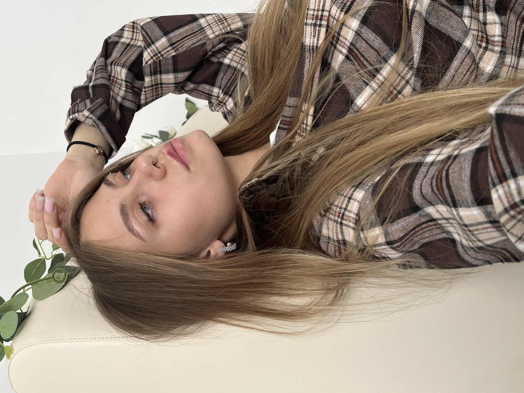 A woman with long blonde hair and fair skin is lying on a light-colored surface. She is wearing a brown plaid shirt and has a nose ring and earrings. Her left hand is raised towards her face. The location is not discernible, and there is no evidence to suggest the image was taken in Фоки, Russia. There are some green leaves and white flowers in the image.