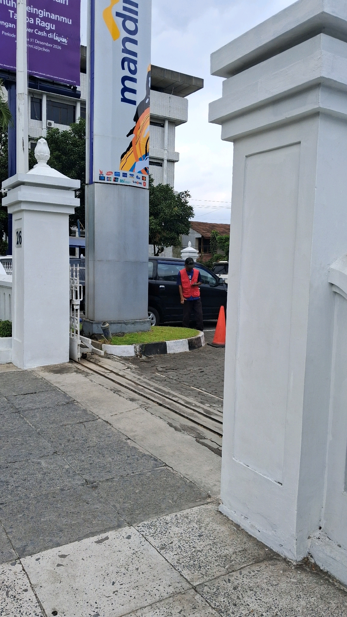 The image shows the entrance to a bank in Yogyakarta, Indonesia. A person wearing a red vest is standing by a black car. The bank has a tall sign with the logo "Mandiri". The entrance is guarded by two white pillars. The ground is paved with cobblestones and tiles. Trees and buildings are visible in the background.