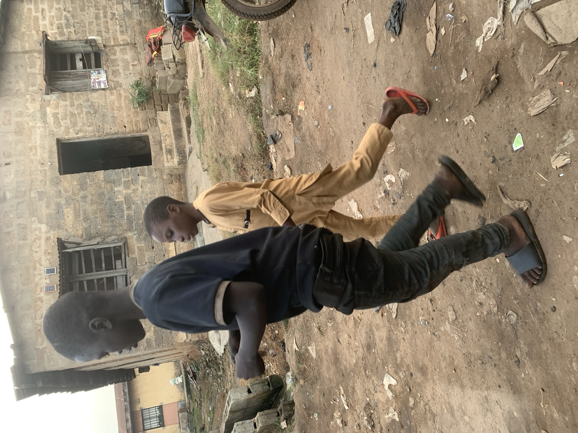 In the city of Shagamu, Nigeria, two young boys walk side-by-side on a dirt path. The boy on the left, in a dark blue t-shirt and black pants, walks with his arms slightly bent and hands clenched. He wears dark grey sandals. The boy on the right, dressed in a tan, loose-fitting garment, walks a few paces behind and to the right of the first boy, wearing red flip-flops. They are in an outdoor, undeveloped area with scattered debris on the ground, including plastic bags, paper, and fabric scraps. Behind them, a weathered, two-story building with a rough brick facade, small windows, and an open doorway stands under a cloudy sky. A portion of a motorcycle is visible on the right edge of the frame, suggesting a semi-urban or rural setting.