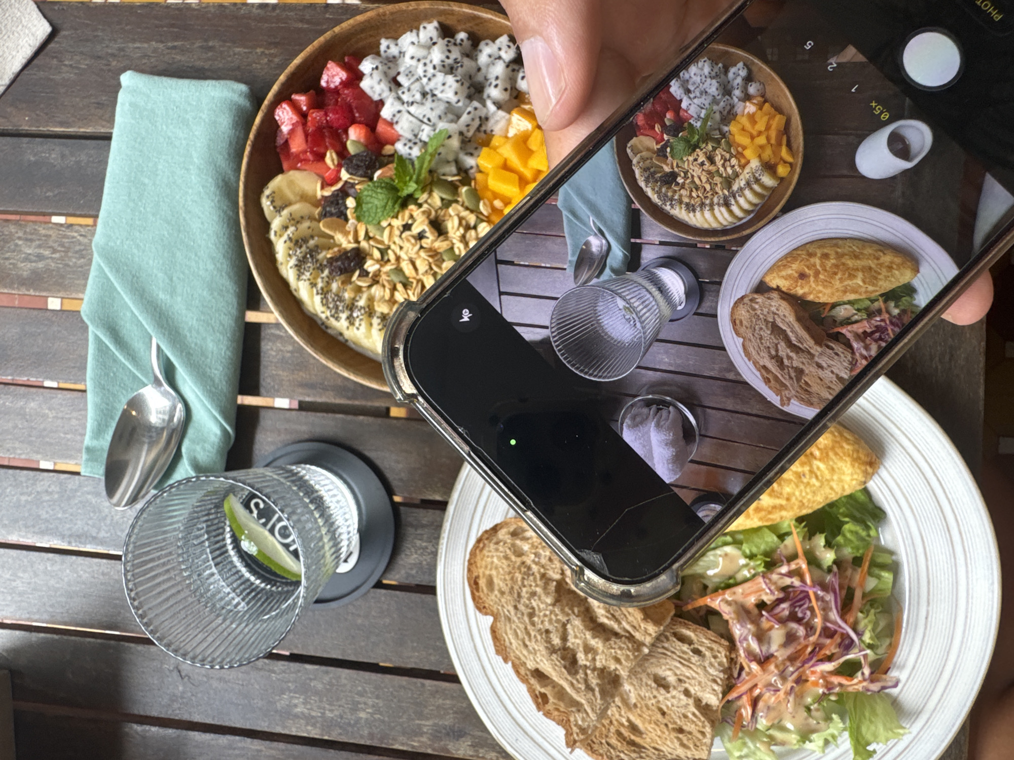 The image captures a dining scene, likely in a restaurant in Đà Nẵng, Vietnam. The scene is shot from an overhead perspective, and the composition consists of two plates, a glass of drink, and a hand holding a smartphone taking a picture of the scene. One plate contains a colorful fruit bowl, with ingredients such as strawberries, dragon fruit, mango, bananas, and granola. The other plate contains what appears to be an omelet with toast and salad. There is a green napkin and a spoon next to the fruit bowl. The glass is filled with a clear liquid, possibly water, with a slice of lime. The background consists of a wooden table with visible grain and textures. The smartphone in the foreground has its camera pointed at the plates, indicating the act of photographing the food.