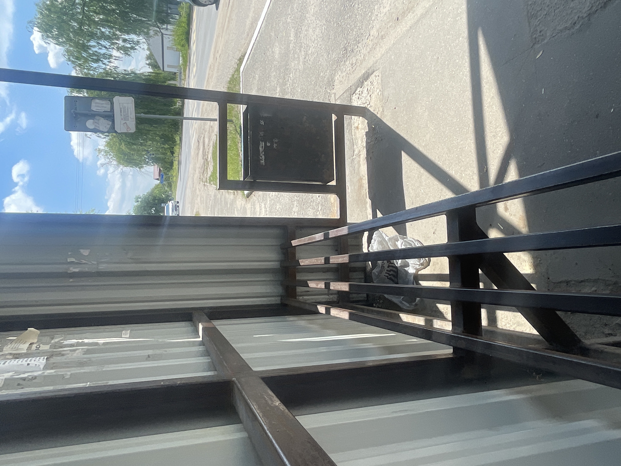 The image shows a bus stop shelter in Мухавецкі сельскі Савет, Belarus. The shelter has a corrugated metal roof and walls, and a metal bench. There is a trash can next to the shelter. The street is visible in the background, with cars and trees. The sky is blue with some clouds. The overall scene is a typical rural bus stop in Belarus on a sunny day.