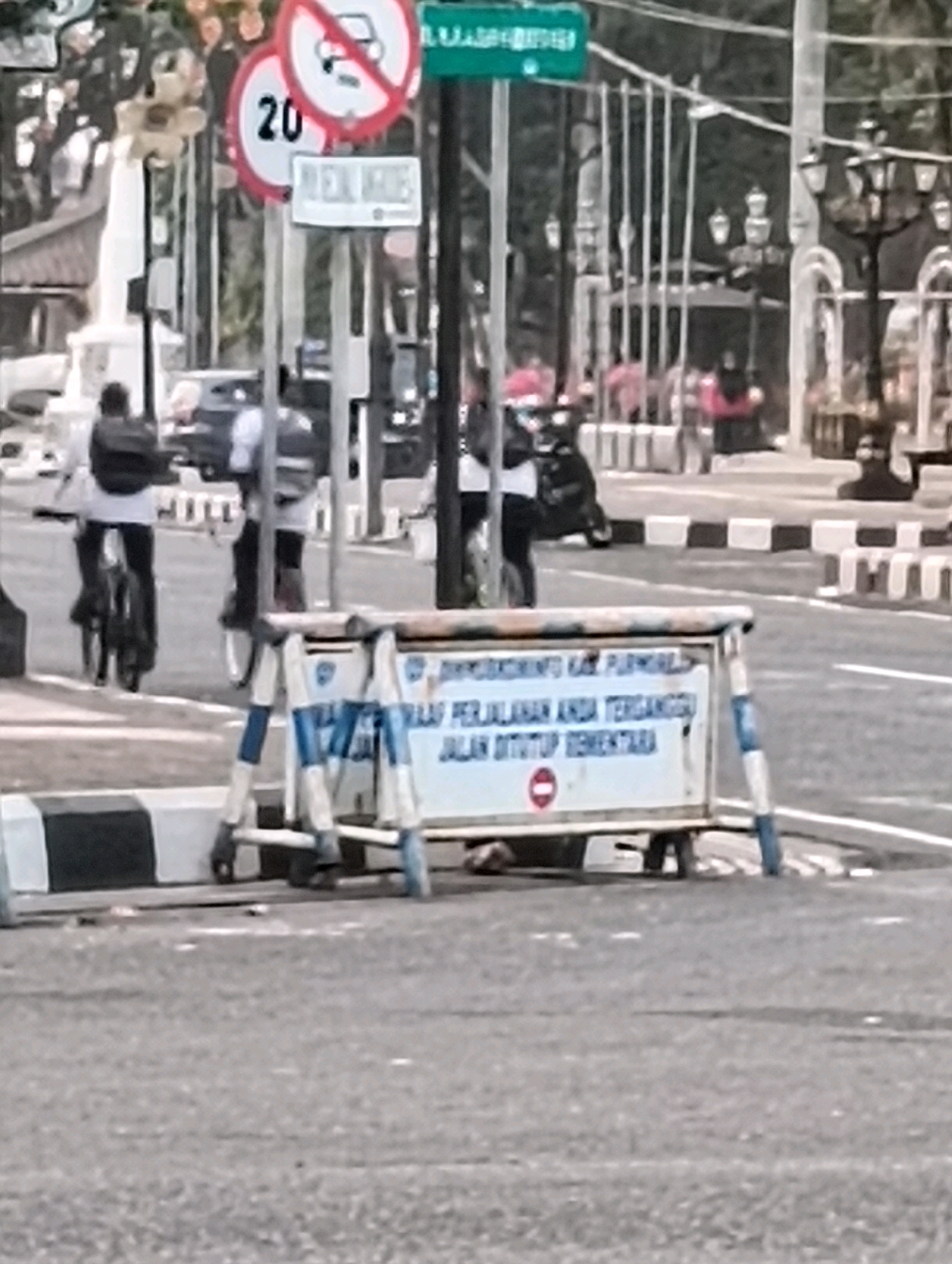 The image shows a street scene in Purworejo, Indonesia. In the foreground, a temporary barrier with a sign is set up, indicating that the road is temporarily closed. The sign reads: "DIMOHON MAAF PERJALANAN ANDA TERGANGGU JALAN DITUTUP SEMENTARA" which translates to "WE APOLOGIZE FOR YOUR INCONVENIENCE, THE ROAD IS TEMPORARILY CLOSED." The barrier is constructed of metal poles and a white panel with blue and white striped tape. In the background, several people are visible, some on bicycles and others walking. Traffic signs are also present, including a "no vehicles" sign and a speed limit sign indicating 20. The environment appears to be urban, with a paved road and sidewalk. Other objects in the background include streetlights and a monument. The overall impression is that of a street closure due to some ongoing event or maintenance work.