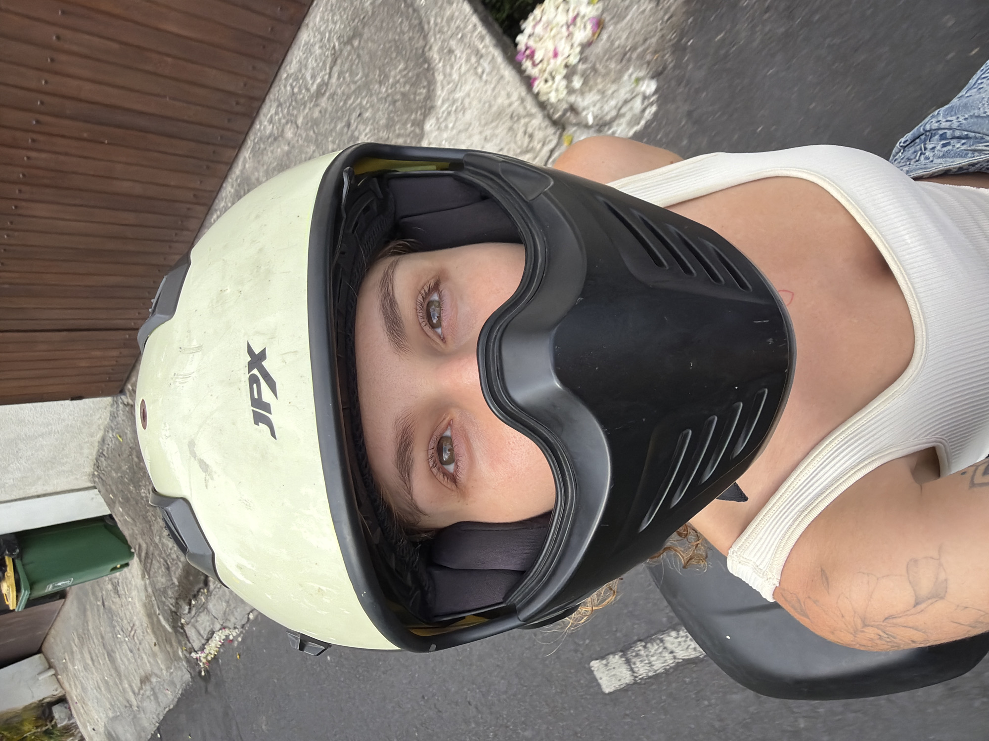 A woman wearing a light yellow JPX brand motorcycle helmet and a white ribbed tank top is taking a selfie while sitting on a motorcycle. Her face is partially obscured by the helmet's black chin guard. The background shows a paved road with white markings, a grey wall, and a wooden fence. A green trash can is visible in the distance. The scene appears to be in a residential or urban area, possibly North Kuta, Indonesia, suggested by the presence of offerings of flowers on the ground, common in Balinese culture.