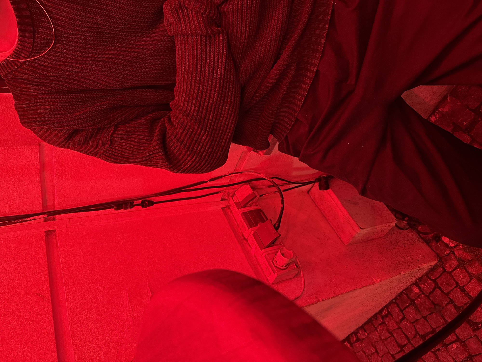 The image shows a person wearing a dark, ribbed sweater and dark pants, standing in front of a red-lit wall. The person has their arms crossed. To the left, a round, reddish object, possibly a table or stool, is partially visible. On the wall, there are electrical outlets with cords plugged in. The ground appears to be paved with cobblestones. The scene is illuminated by a red light, giving a monochromatic red hue to the entire image. The location is identified as Unknown, Unknown.
