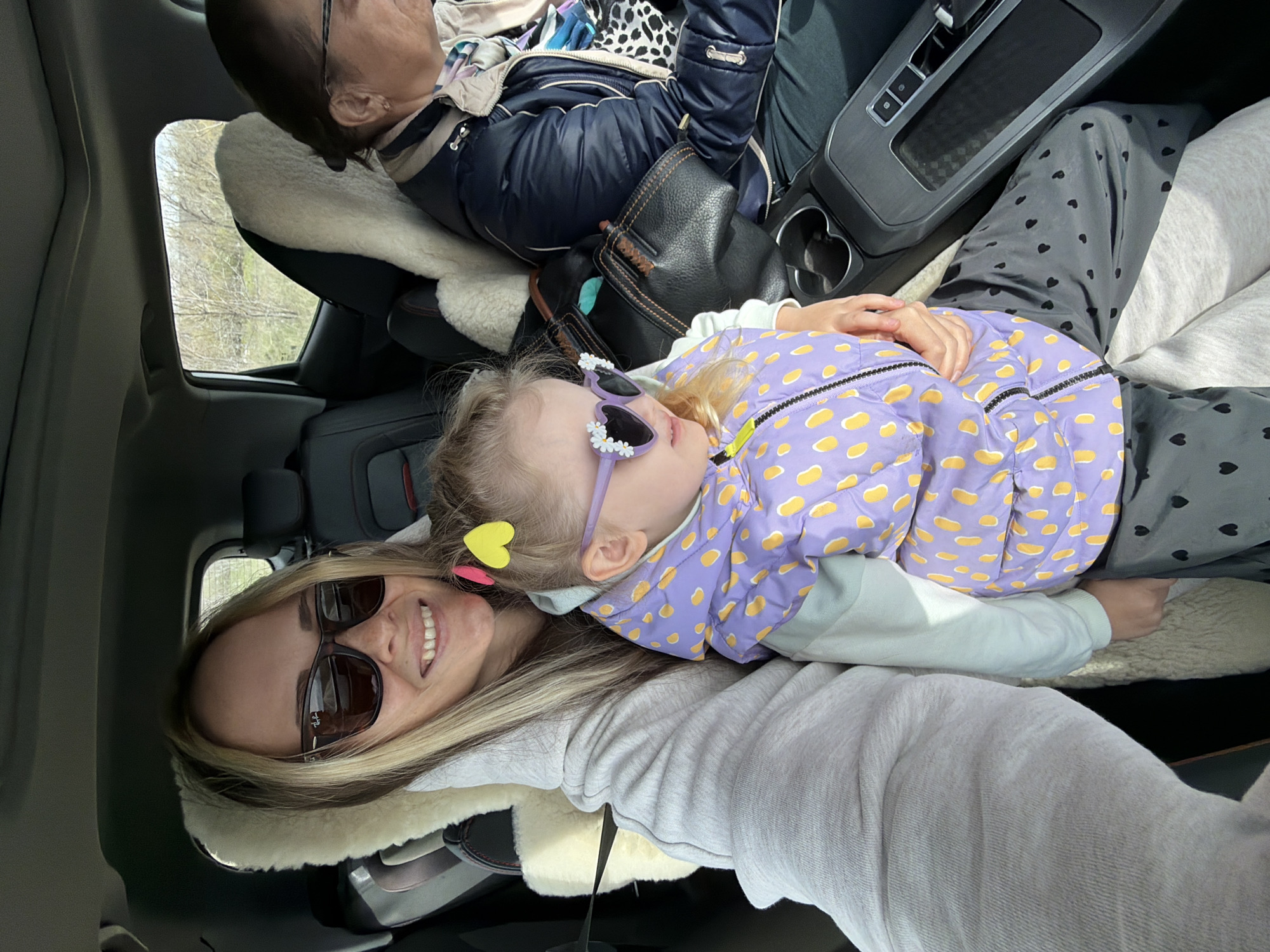 The image shows three people inside a car. A woman in sunglasses is in the foreground smiling, with her head resting on a sheepskin car seat cover. A young girl wearing a purple and yellow patterned jacket and sunglasses is lying down next to her. Behind them, a third person is seated, wearing glasses and a blue jacket. The car's interior is visible, including the center console and a window showing an outdoor scene with trees. The location, Ternovka, Russia, is not visually confirmed in the image.