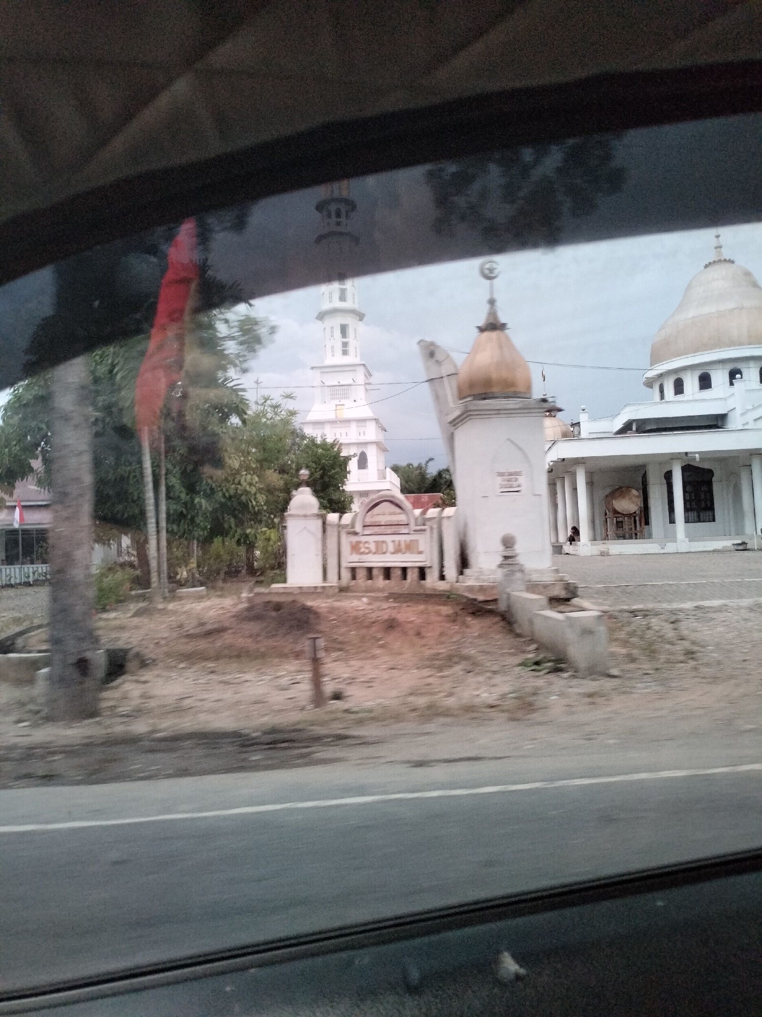 The image shows a mosque and its surroundings, possibly in Lubuk Kasih, Indonesia. The main mosque building has a large white dome and is supported by pillars. Adjacent to it is a white minaret with multiple levels and a pointed top. In the foreground, there is a sign that reads "MESJID JAMIL" in capital letters, indicating it is a mosque. The area around the mosque appears to be paved, with some dirt and sparse vegetation. A few trees are visible, and a red flag is planted on a pole to the left of the mosque. The scene is viewed from a slightly elevated perspective, suggesting it might have been taken from a vehicle. The sky is overcast, indicating a possibly cloudy day.