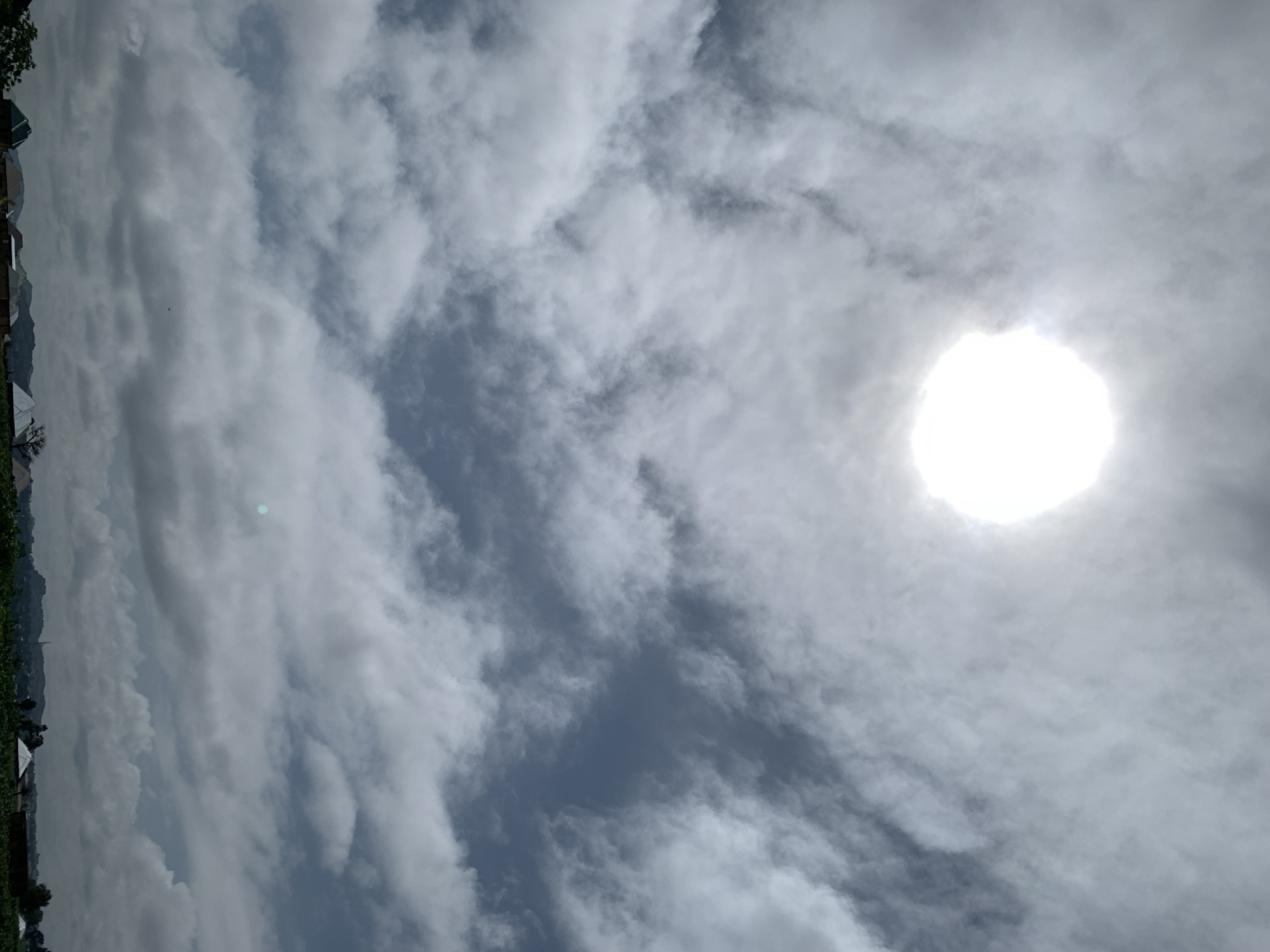 The image captures an overcast sky over Kuru Karama, Nigeria, with the sun partially obscured by thick clouds, creating a bright, diffused halo. The landscape below features a collection of residential buildings with varied roof structures, some appearing to be made of corrugated metal and others of darker materials. A line of low-lying hills or mountains forms the horizon in the distance. The foreground is filled with dense green vegetation, suggesting agricultural activity or natural growth. The overall scene depicts a typical day in the region, characterized by cloudy weather and rural settlement.