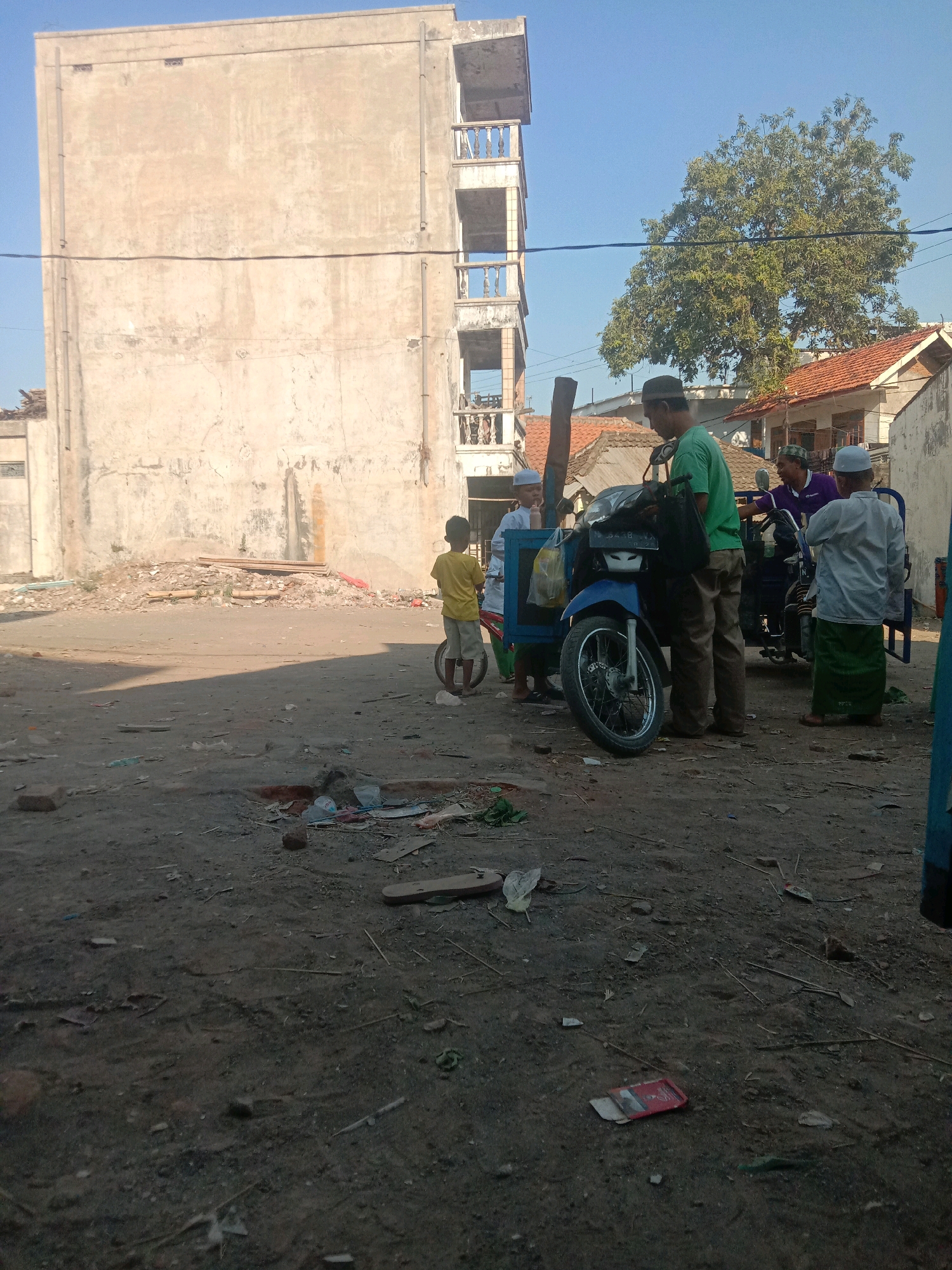 The image depicts an outdoor scene on a sunny day, likely in the city of Sidogiri, Indonesia, given the context. In the foreground, the ground is uneven and dusty, littered with various debris including plastic bags, scattered wood, and a red rectangular object. A young boy in a yellow shirt and light-colored shorts stands near a blue motorcycle that is parked on the dirt. Behind the motorcycle, several men are gathered. One man in a green shirt and brown pants appears to be near the motorcycle, while another man in a purple shirt is partially visible. Two other individuals, one in a white shirt and another in a white cap and shirt, are also present.
In the background, a large, weathered building with multiple balconies stands prominently. To the right of the building, a smaller house with an orange tiled roof is visible, along with a leafy green tree. The overall impression is that of a somewhat unkempt urban or semi-urban environment.