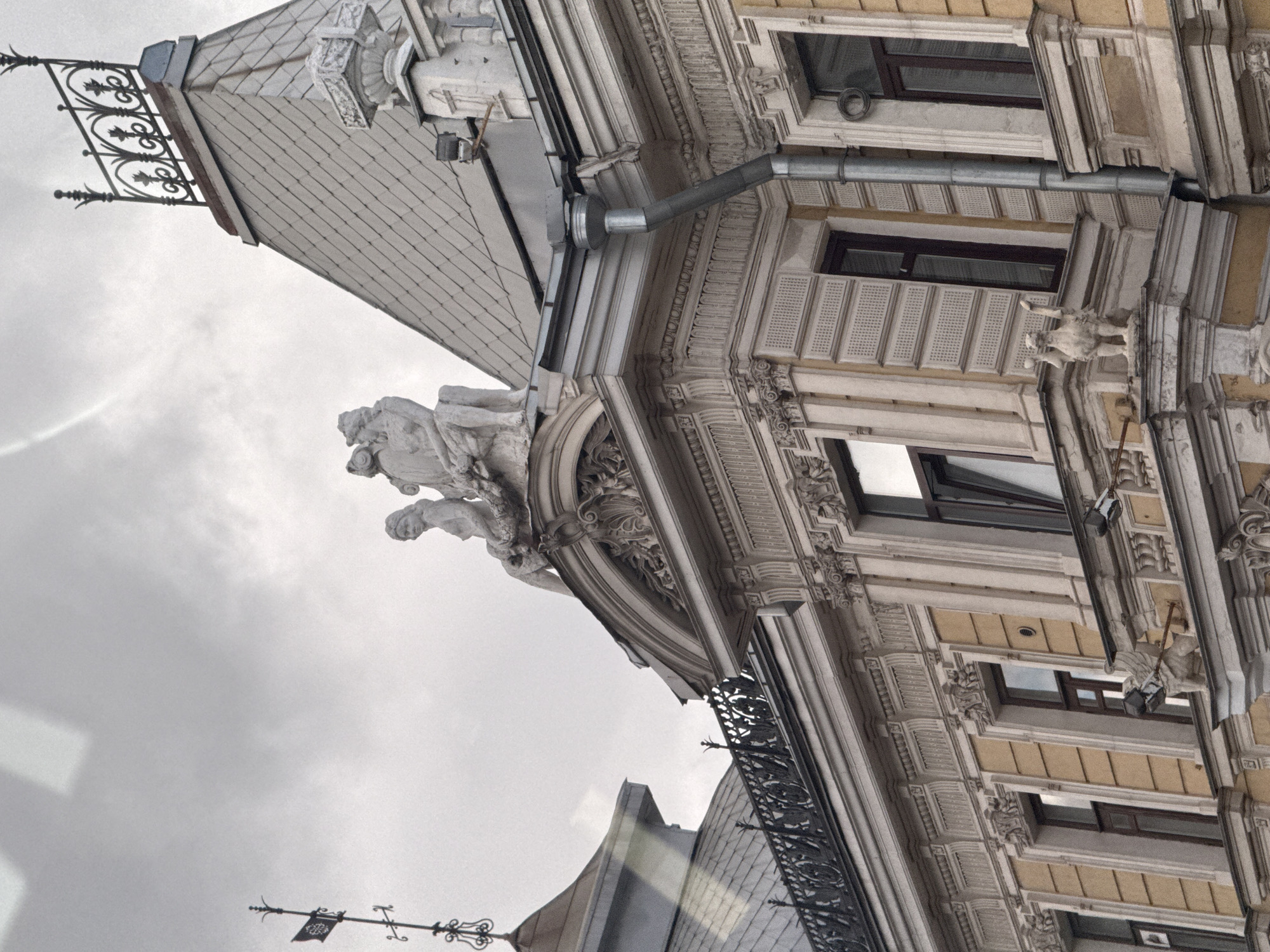 This image shows an ornate building exterior with classical architectural elements. At the top of the building, there are several stone statues, including a group of figures seated on a ledge and a mythical creature with wings. Decorative ironwork adorns the roofline. The building has multiple windows with dark frames, and the facade is a pale yellow color with intricate white trim and carvings. A downspout is visible on the right side of the building. The sky is overcast and grey. There are no people or animals visible in the image. The scene appears to be in a city environment, likely with a historical architectural style. The location in Moscow, Russia is not discernable from the image.