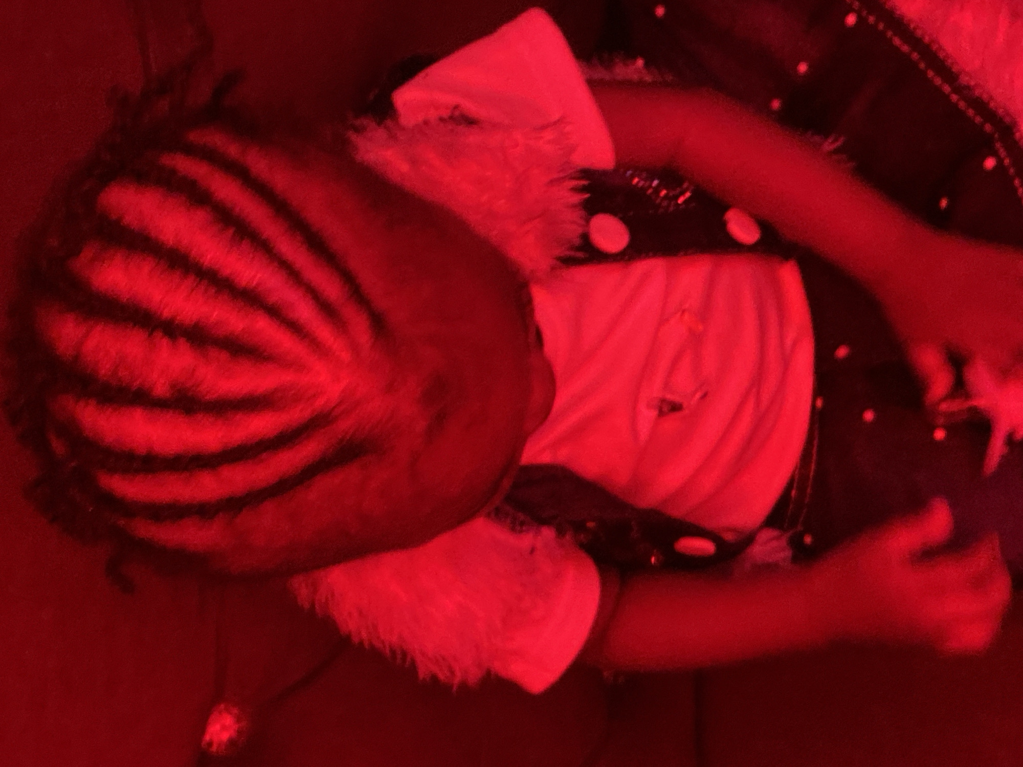 A child is depicted in a red-lit environment, possibly a room. The child's hair is styled in distinct cornrows that are illuminated by the red light. They are wearing a white t-shirt and a dark vest adorned with circular sequins. The child's left arm is bent, with their hand visible. The background is dark, with some out-of-focus red lights creating bokeh effects. There is no indication of the city of Abakaliki, Nigeria, or any specific urban setting within the image. The image appears to be a portrait of the child.