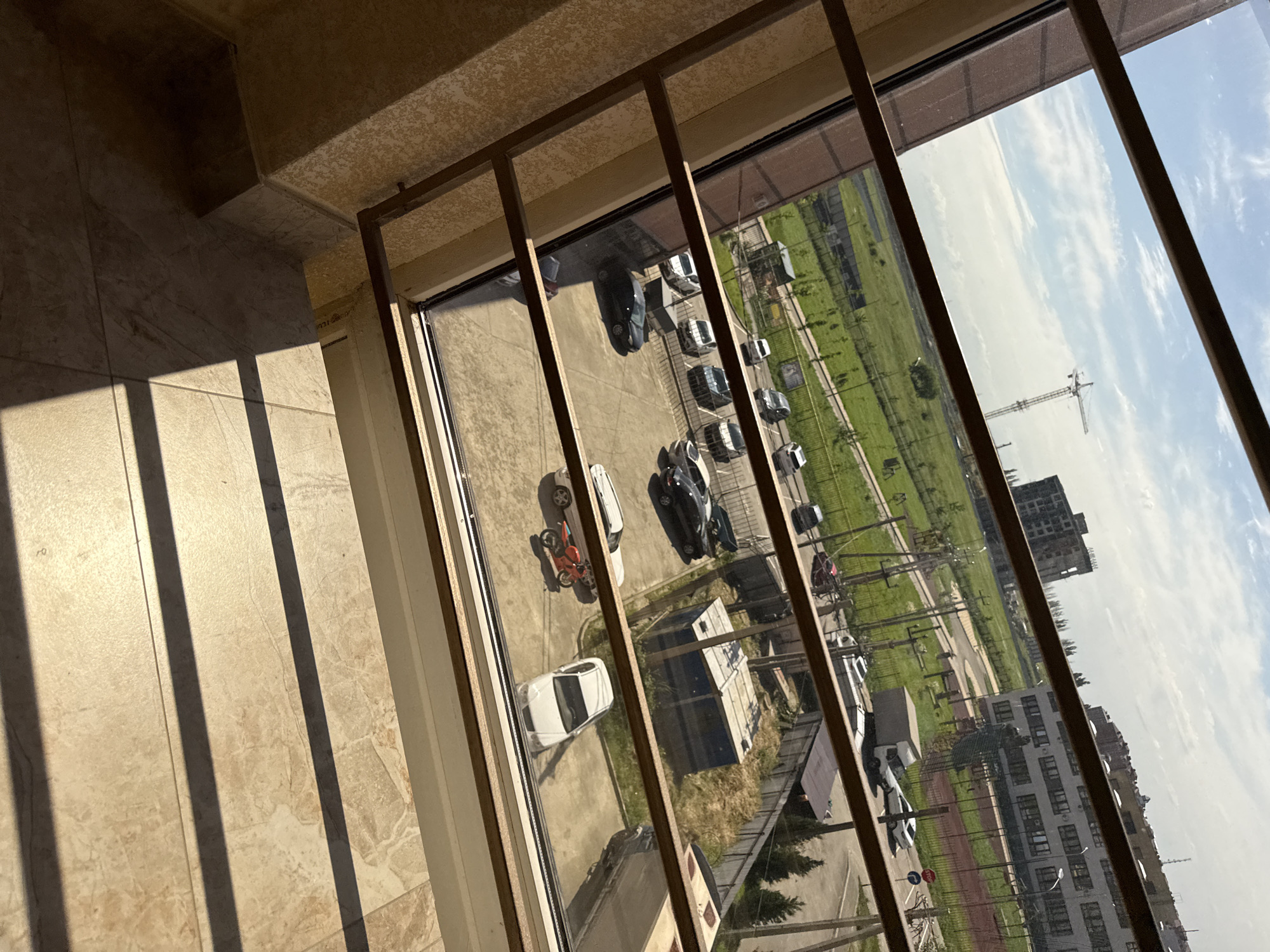The image, captured from an indoor perspective looking out a window, depicts a slanted view of an outdoor scene under bright daylight. Inside, parallel shadows from a window frame or metal bars are cast across a light-colored, possibly tiled or patterned floor.
Beyond the window, a diverse environment unfolds. Multiple vehicles, including cars (sedans and SUVs, some black, some white) and a red motorcycle, are visible in a parking area or on a road. Beyond the vehicles, a large expanse of green field stretches out, dotted with trees. In the distance, several multi-story buildings are present, one of which appears to be an unfinished construction site with a skeletal structure, suggesting ongoing development. A large construction crane stands tall against the horizon. The sky is bright blue with scattered white clouds.
The strong, distinct shadows cast by the window bars on the floor suggest it is daytime, likely morning or late afternoon. The weather appears clear and sunny. There is no specific activity overtly taking place beyond the static presence of vehicles and buildings. No clearly legible text is visible in the image.