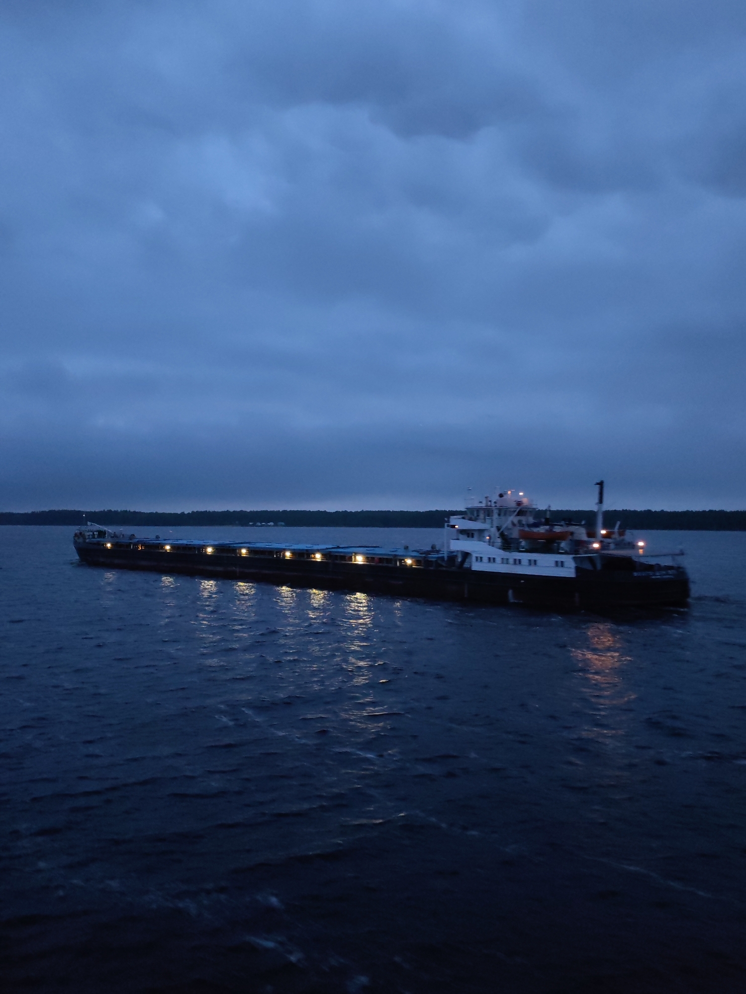 A long, dark barge with a multi-tiered white superstructure is traveling on a body of water under an overcast sky. The barge has numerous lights illuminated along its length, reflecting in the water. Dark vegetation appears on the shoreline in the distance. The photo was taken in Voznesenye, Russia.
