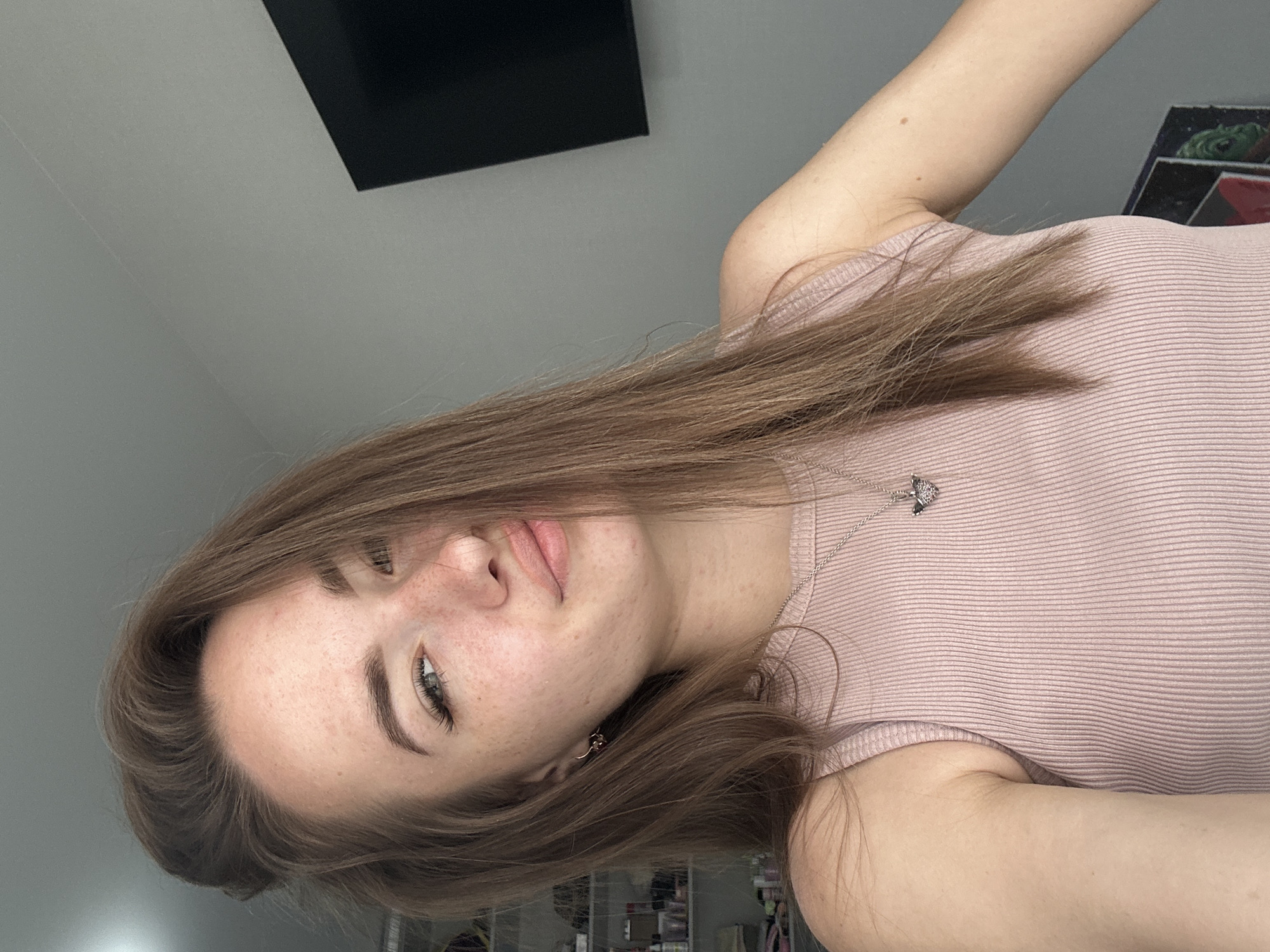A young woman with brown hair and freckles is taking a selfie indoors. She is wearing a pink ribbed tank top and a silver necklace with a heart pendant. Her hair is partially covering her face. In the background, there is a gray wall with a black television mounted on it. There are also shelves with various items on them.