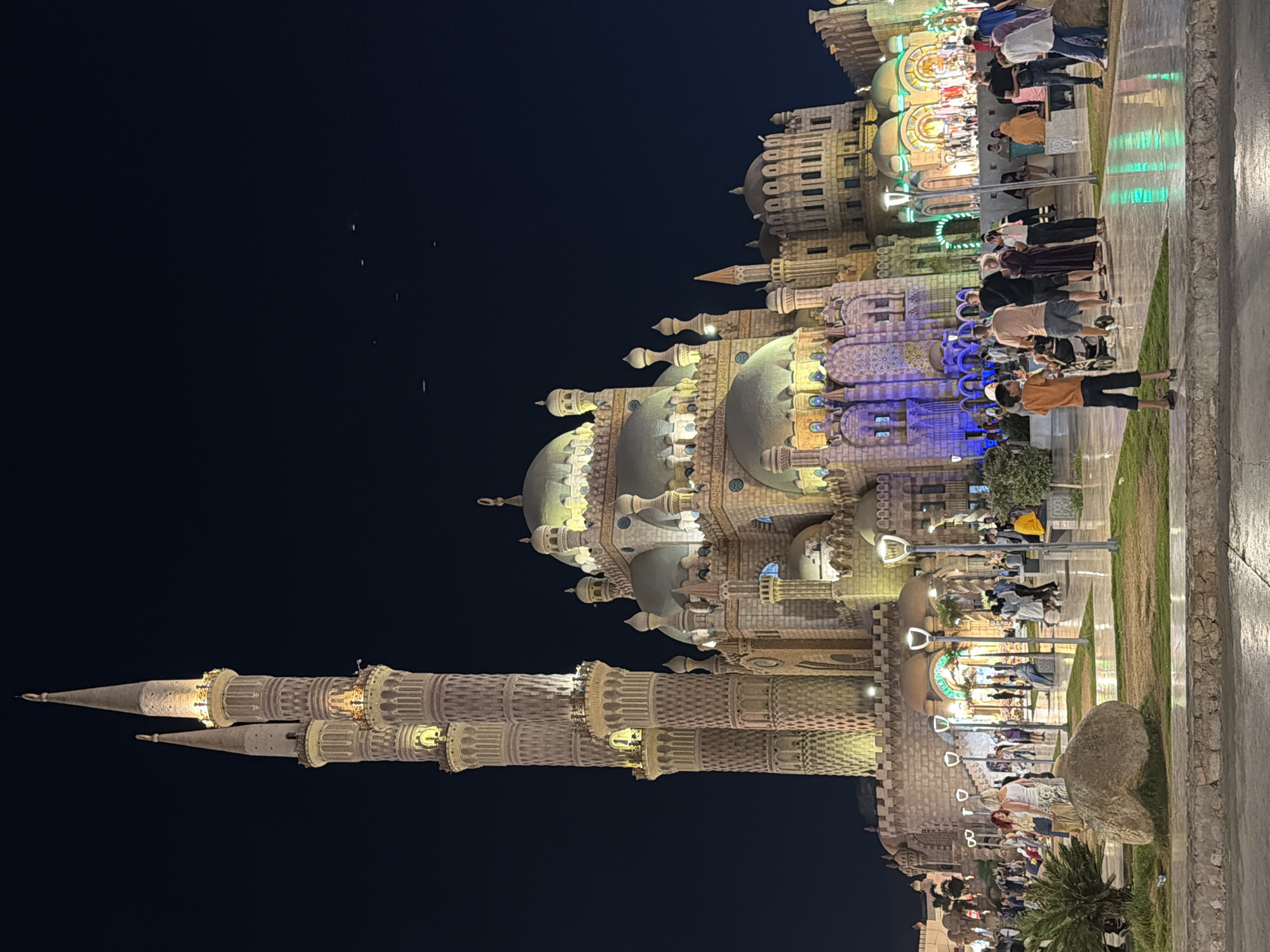 The image depicts the Al-Sahaba Mosque in Sharm El Sheikh, Egypt, at night. The mosque is a prominent landmark with its distinctive architecture, featuring multiple domes and tall, ornate minarets illuminated by warm lighting. The scene is bustling with people of various ages and backgrounds, some walking, some standing in groups, and others sitting. The ground appears to be wet, reflecting the colorful lights of the mosque and surrounding lampposts, creating a vibrant and lively atmosphere. The sky is dark, indicating it is nighttime, with a few faint streaks of light, possibly from aircraft or distant lights. The overall impression is of a lively public space surrounding a significant religious structure.