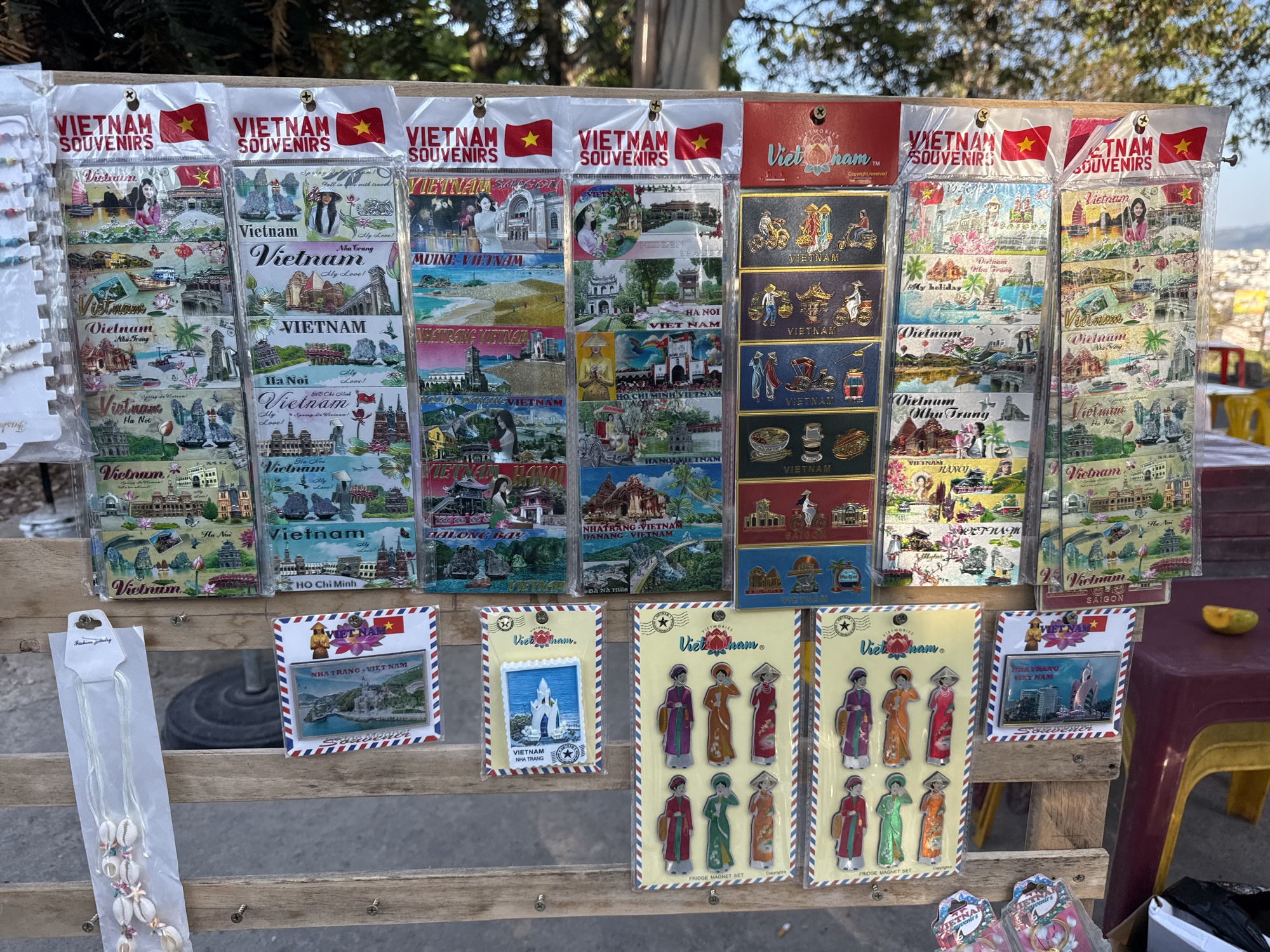 An outdoor souvenir stand in Tay Nha Trang Ward, Vietnam, displays various Vietnam-themed magnets. These include depictions of landscapes, cityscapes, and cultural icons. They are packaged in plastic bags and hung on a wooden rack, including magnets featuring traditional Vietnamese clothing. Additionally, there are shell necklaces and souvenir keychains visible. The background includes trees, a building, and a partially visible cityscape. A yellow chair with a red seat sits nearby.
