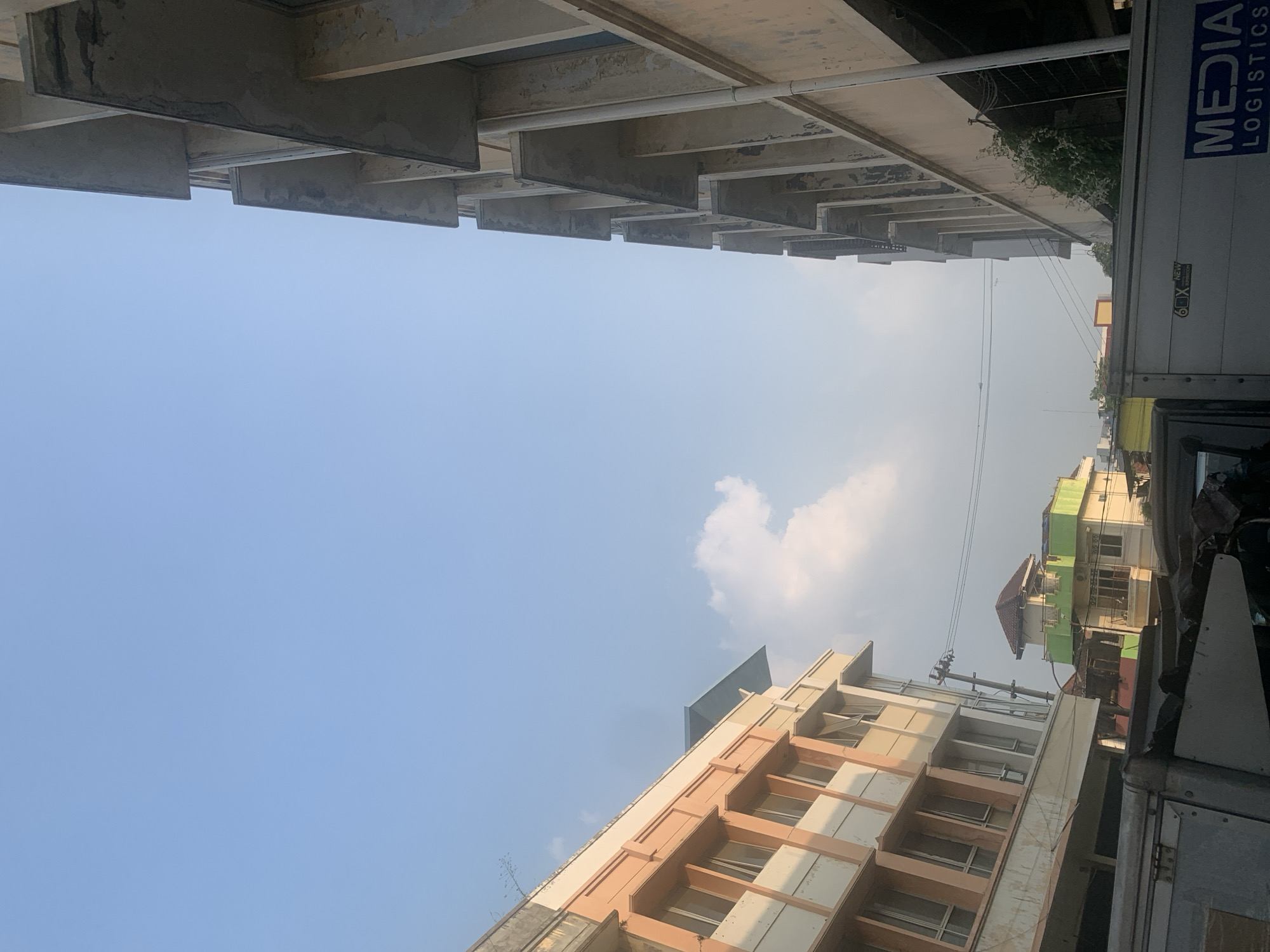 The image shows an outdoor urban scene with buildings and a truck in the foreground. On the left side of the image is a pale peach and cream colored building with multiple floors and windows. The building on the right is a multi-story structure with a light beige facade, characterized by protruding concrete elements that create a series of overhangs or balconies. The sky is clear blue with a few scattered clouds. In the background, partially obscured by the buildings, is another structure with a green and white facade and a brown roof. A portion of a white truck with "MEDIA LOGISTICS" written on its side is visible in the lower part of the image. There are power lines and a utility pole visible between the buildings. The overall scene suggests a commercial or residential area. The presence of the "MEDIA LOGISTICS" truck suggests this location is within a business or industrial area of Palembang, Indonesia.