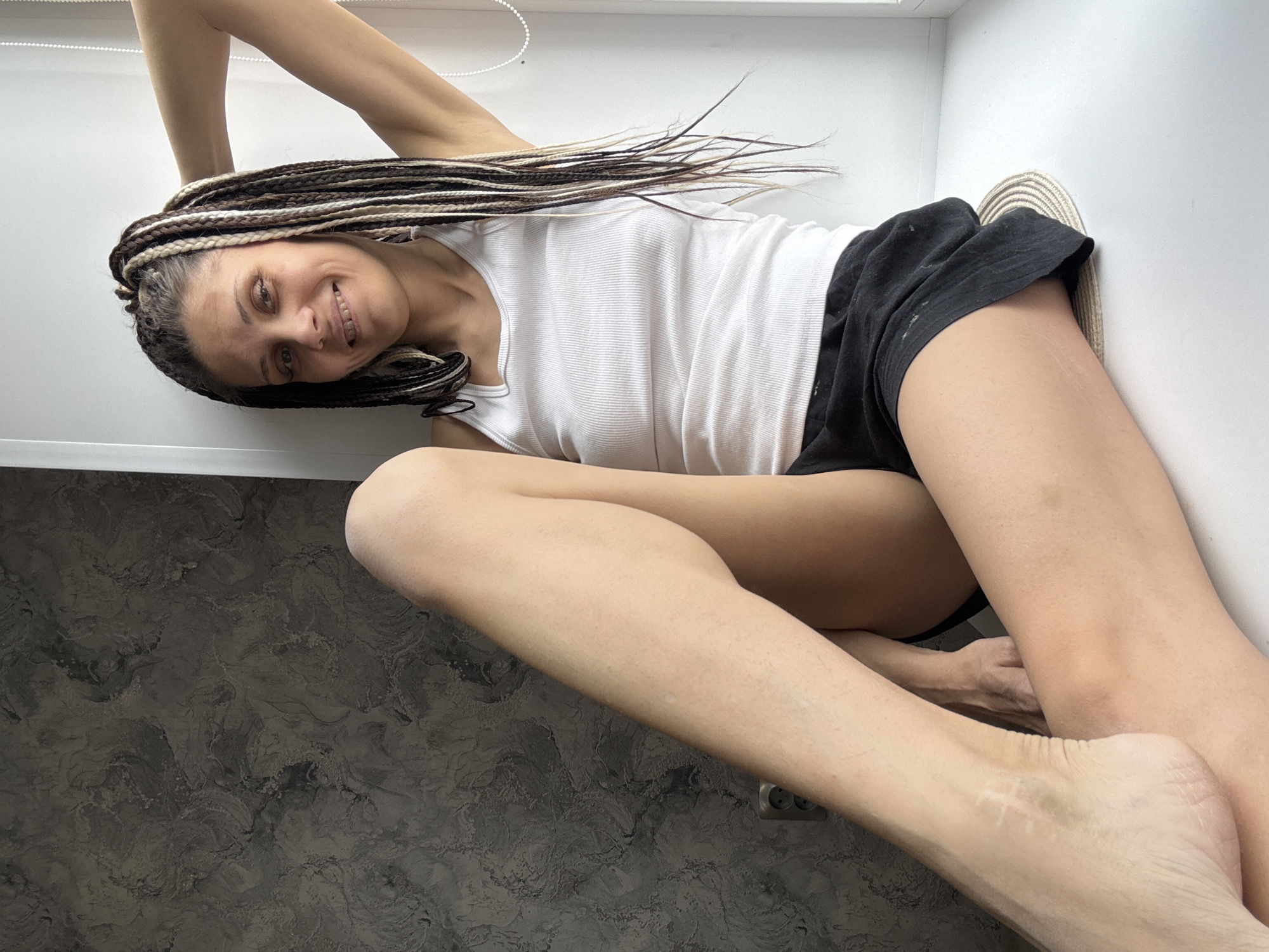 A woman lies in an Unknown, Unknown room. She is wearing a white tank top and black shorts. She is smiling. The room has a gray countertop, white walls, and a white blind cord.