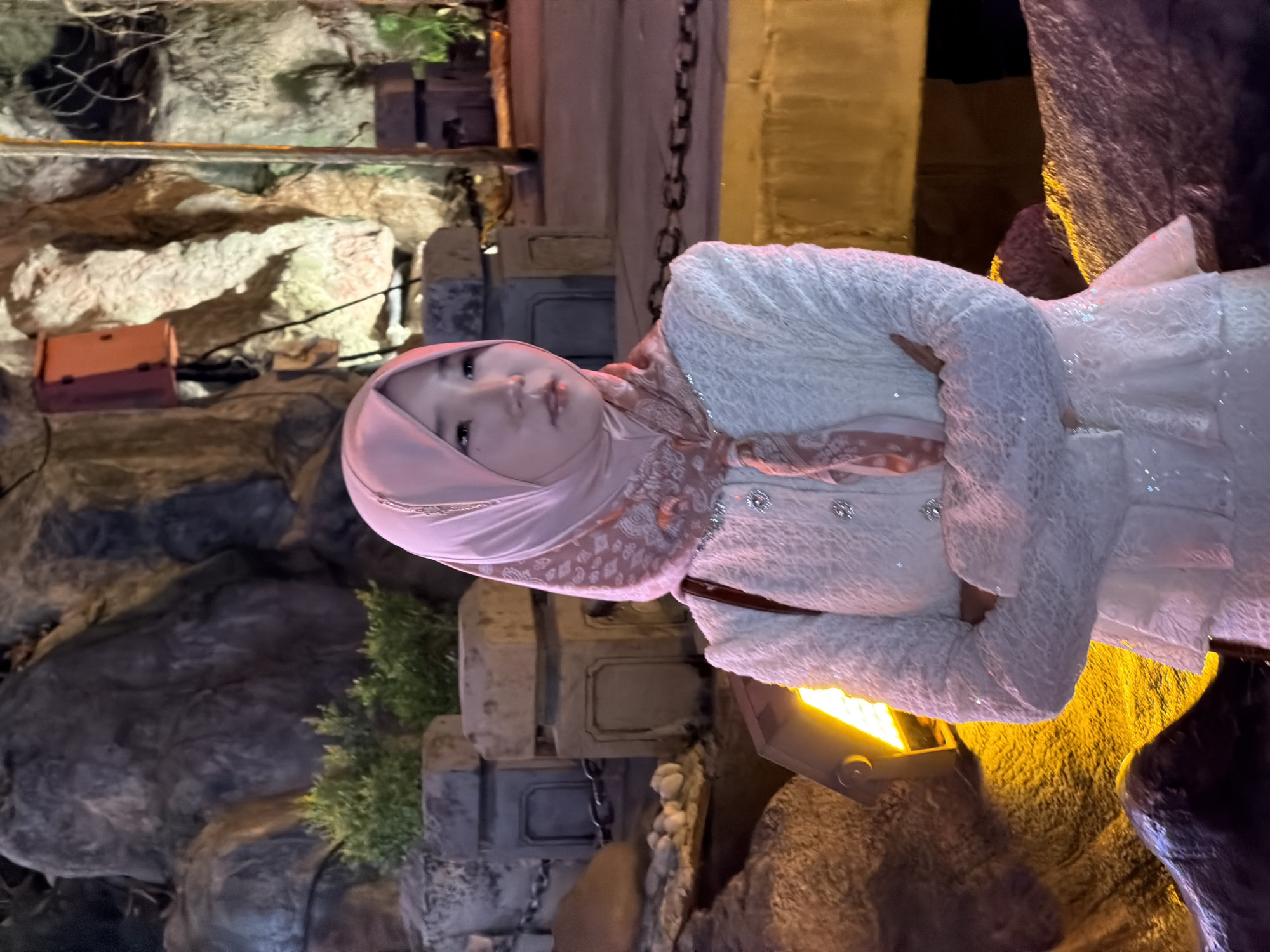 A young woman wearing a pink hijab and a white lace dress stands with her arms crossed in front of a rocky, cave-like background. The scene appears to be outdoors at night, with artificial lighting casting warm glows on the rocks and foliage. In the background, there are stone structures and what looks like a chain barrier. The woman is looking off to the side with a neutral expression. The location is identified as Yuzhong District, China.
