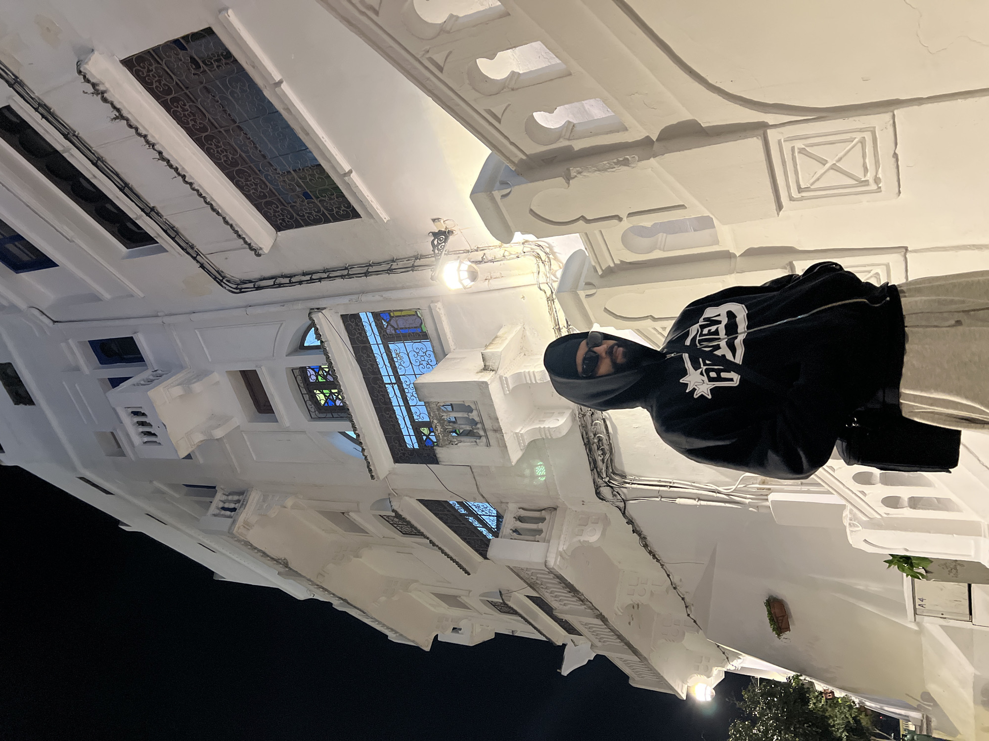 The scene depicts a male subject standing at night in front of a multi-story, whitewashed building characteristic of Tangier, Morocco. The subject, viewed from the chest up, wears a black hooded sweatshirt with a white graphic print, dark sunglasses, and light-colored sweatpants. A black shoulder bag is worn, and his hands are placed in his pockets. He has a visible beard. The prominent building behind him features ornate architectural details including multiple balconies with white balustrades, windows fitted with decorative dark wrought iron grilles, and internally illuminated colored glass panels displaying blue, green, and yellow light. A wall-mounted lamp with an opaque globe illuminates the area near a white staircase or railing, which incorporates stylized arch-shaped cutouts. Exposed electrical conduits are visible on the building's exterior. A small brown planter with vegetation and a white utility box labeled "A4" are present on the lower left portion of the facade.