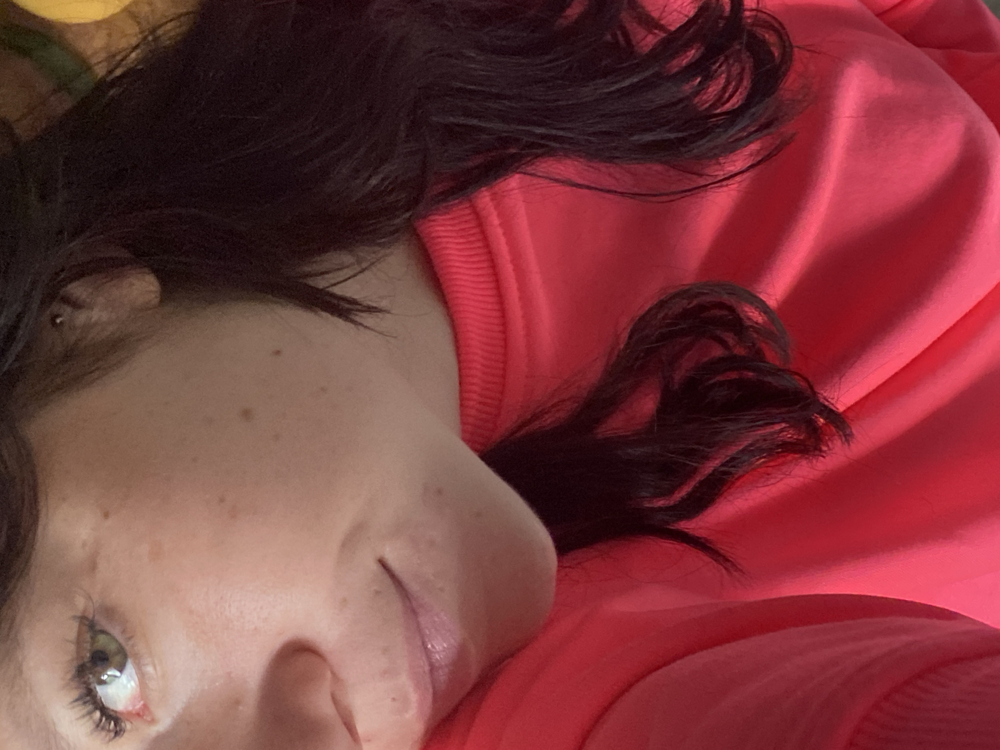 A close-up, medium shot captures a portion of a person's face and upper body, oriented slightly to the left. The individual has light skin with freckles, a green eye with long eyelashes, and dark, wavy hair that drapes over their shoulder. They are wearing a bright pink sweatshirt with a ribbed neckline. The lighting is soft, creating a gentle illumination. The background is out of focus, with hints of yellow and green that suggest an indoor setting, possibly a home, with some decorative elements. The overall tone of the image is casual and intimate. The location in Chaikovsky, Russia, is not discernible from the visual information provided in the image.