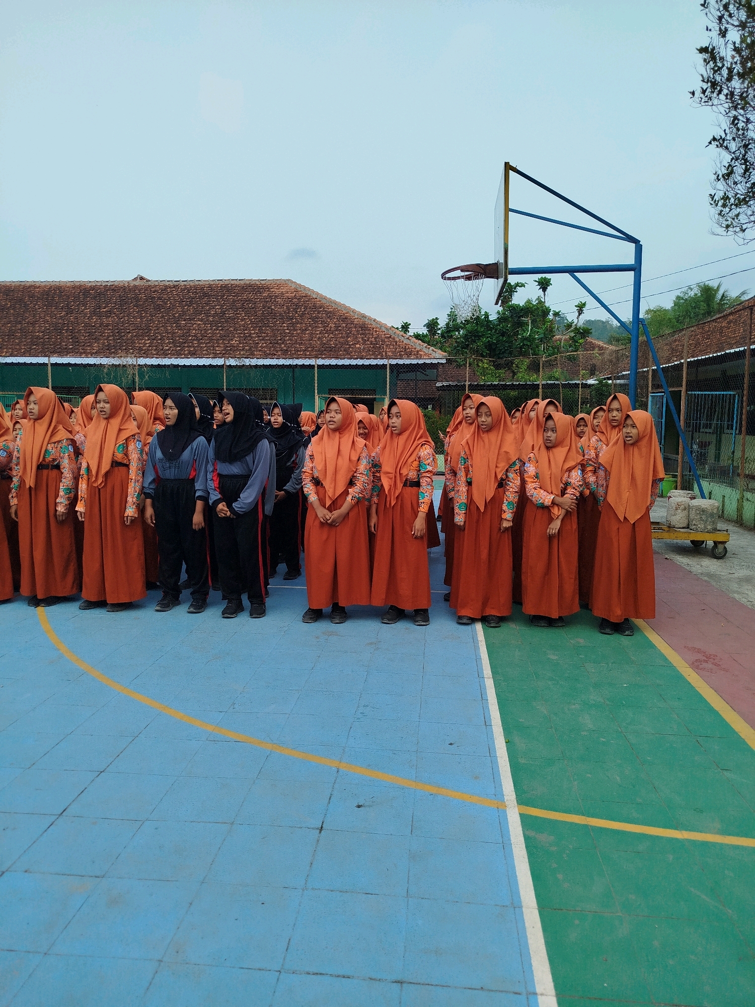 In Karangsambung, Indonesia, a group of young women wearing orange hijabs and patterned shirts stand in formation on a blue and green tiled basketball court. Their attire suggests a school uniform. Behind them, a building with a red tiled roof and green trim is visible, along with a basketball hoop and a fence. The sky is overcast and the lighting is diffused, indicating it might be early morning or late afternoon.