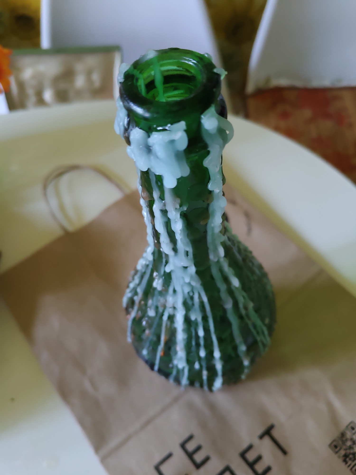 The image shows a close-up of a green glass bottle candle with white wax drips. The candle is sitting on a light-colored surface, possibly a table, with a brown paper bag in the background. The bag has text printed on it, which appears to be "FFCE EET" and a QR code. The background is blurred, but suggests an indoor setting with some furniture and a hint of a window. There are no people visible in the image, and no specific location in Rostov-on-Don can be identified.