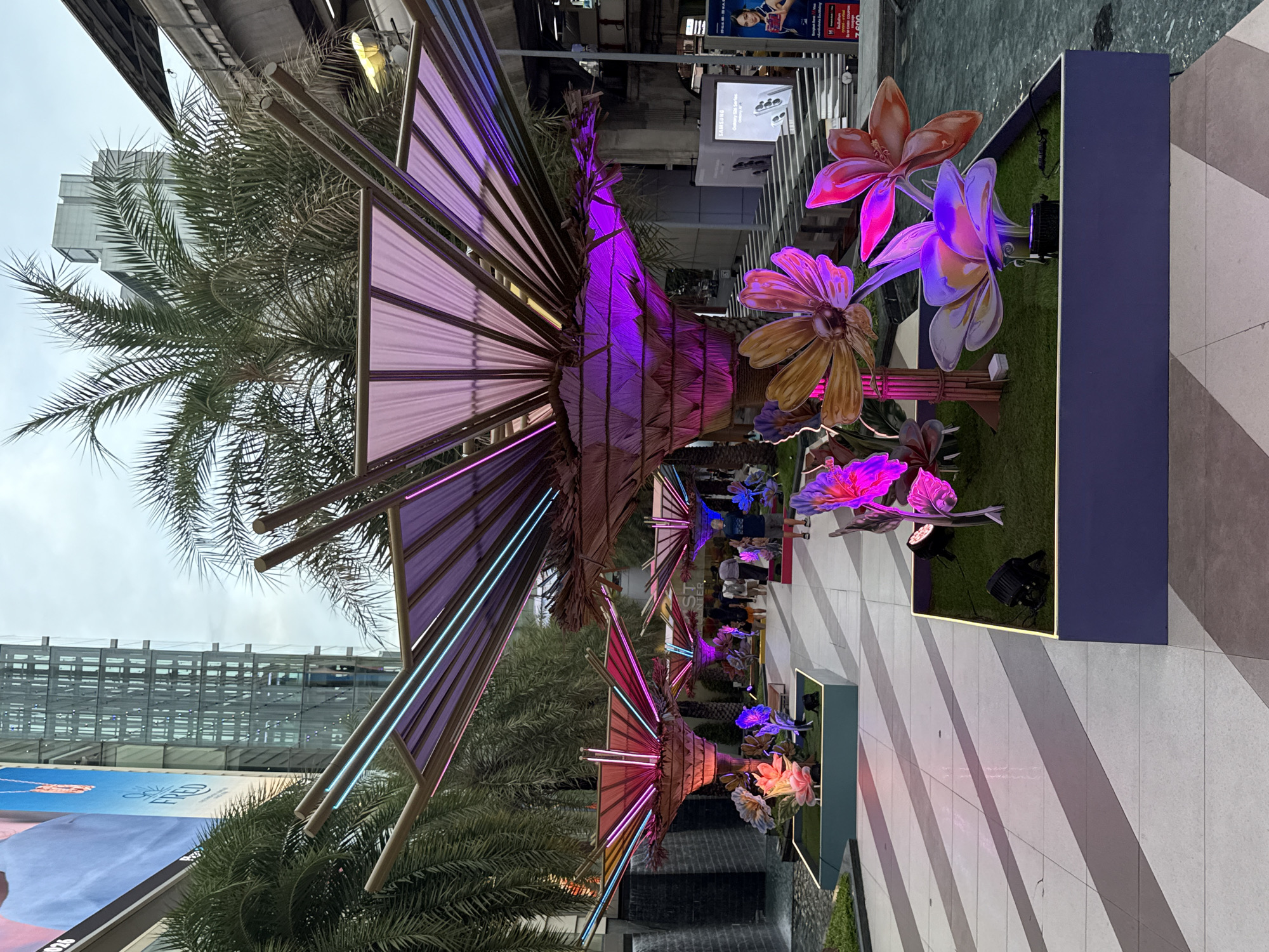This outdoor, eye-level, daytime shot depicts a pedestrian walkway in Bangkok, Thailand, featuring a series of illuminated floral installations and palm trees. The walkway is paved with light-colored tiles in a striped pattern. Overhead, a structure resembling a stylized palm frond is constructed with metal beams and translucent panels, illuminated with purple and pink neon lights.
Below, rectangular planters are filled with artificial turf and oversized, brightly colored artificial flowers illuminated with spotlights. People are visible walking in the distance, suggesting a busy urban environment. The architecture in the background includes modern buildings and elevated transit infrastructure. Advertisements and billboards are visible, contributing to the urban backdrop. A small water feature, possibly a fountain or a decorative pool, is also present along the walkway.