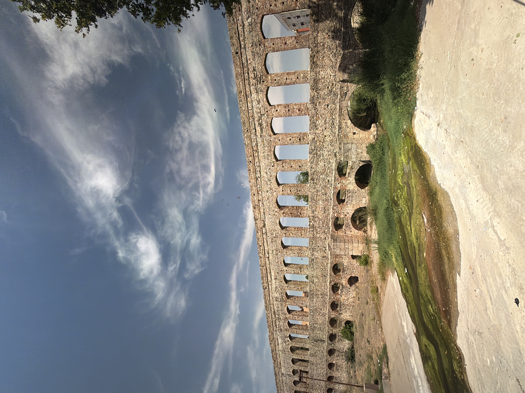 The image depicts a wide shot of an ancient stone aqueduct under a partly cloudy sky. The aqueduct is constructed from rough-hewn stone blocks and features a long series of arched openings on its upper level, through which the sky is visible. Below these, a second row of smaller, darker arches are visible, some of which appear to be submerged in water or overgrown with vegetation. The foreground shows a dry, textured ground surface, possibly concrete or compacted earth, with a patch of greenish, algae-filled water flowing diagonally across it. Lush green grass and reeds grow along the banks of this water feature and around the base of the aqueduct. In the distance, through some of the aqueduct's arches, hints of modern buildings can be seen, suggesting an urban or semi-urban setting. A few small dark shapes, possibly birds, are visible in the sky and near the aqueduct. The overall impression is one of historical ruin integrated into a natural environment, with a stark contrast between the weathered stone structure and the vibrant greenery and water. This specific aqueduct is the Acueducto de los Milagros, located in Mérida, Spain.
